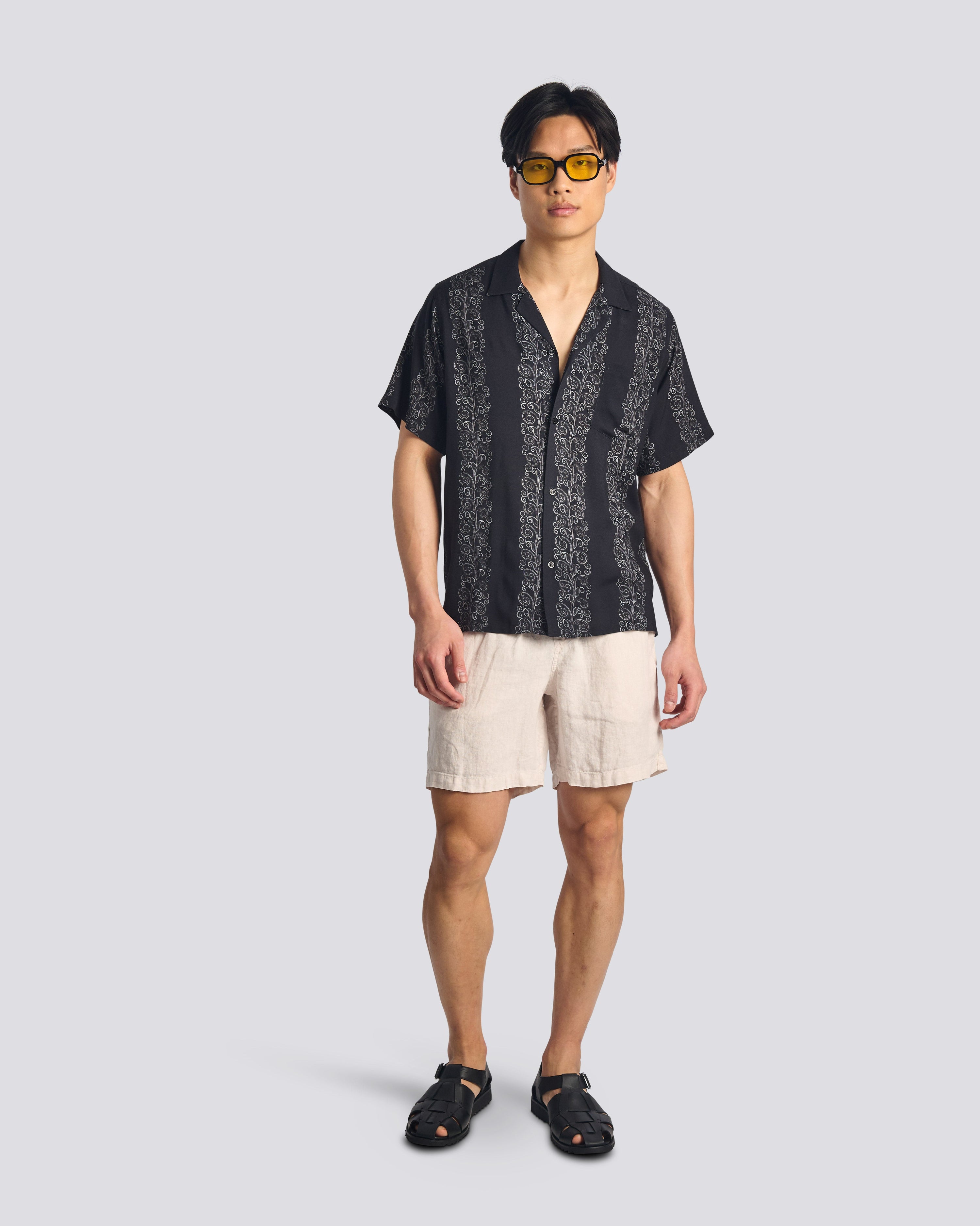 A model wearing the Bather Men's Black Fiesta Camp Shirt, styled with cream trousers. The image showcases the shirt's relaxed fit and the lightweight black rayon fabric with a subtle vertical "Fiesta" stripe pattern.