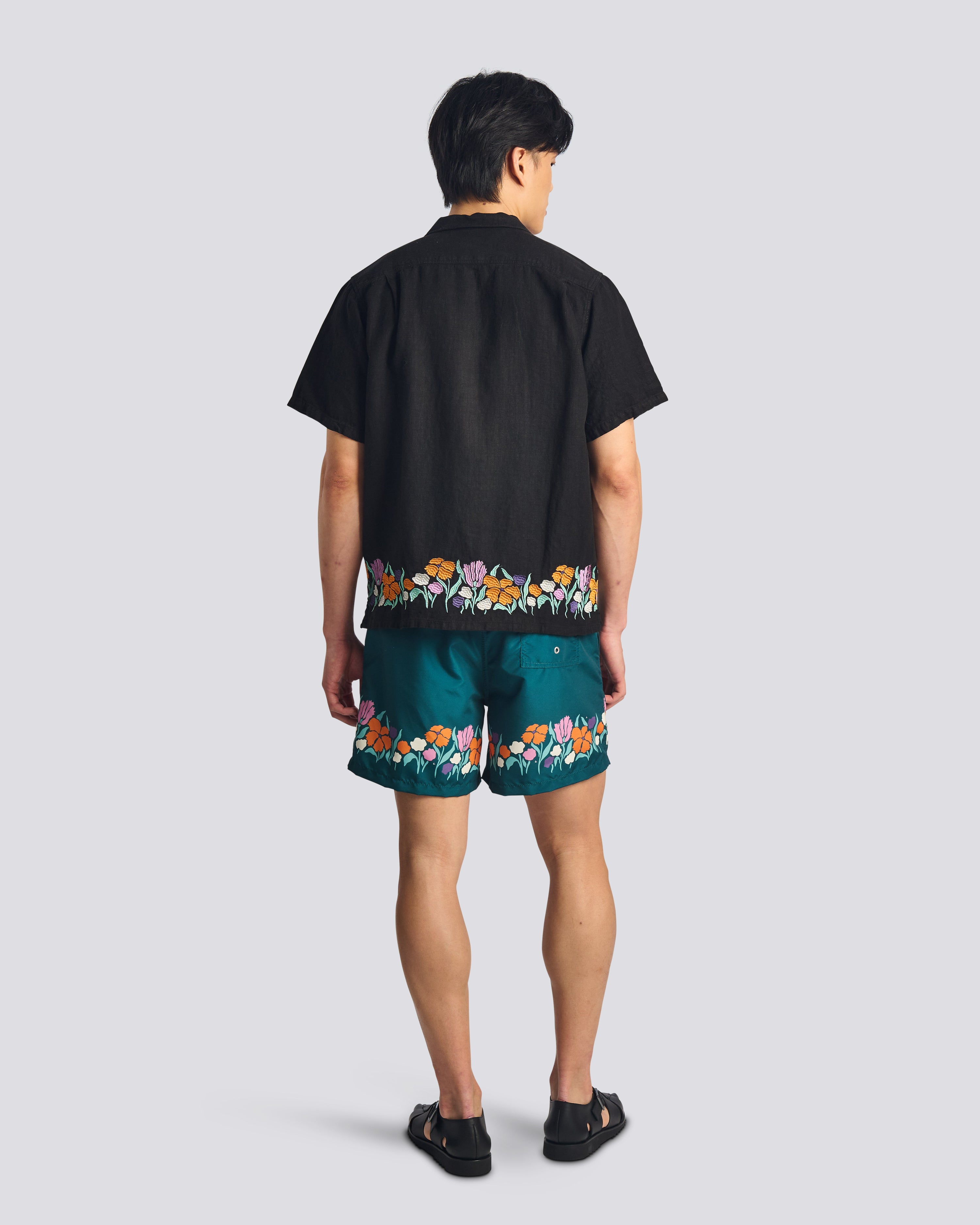 Back view of a model wearing the Bather Men's Black Lawn Linen Camp Shirt, highlighting the black linen fabric and the vibrant floral embroidery wrapping around the hem, styled with matching swim trunks and black sandals.