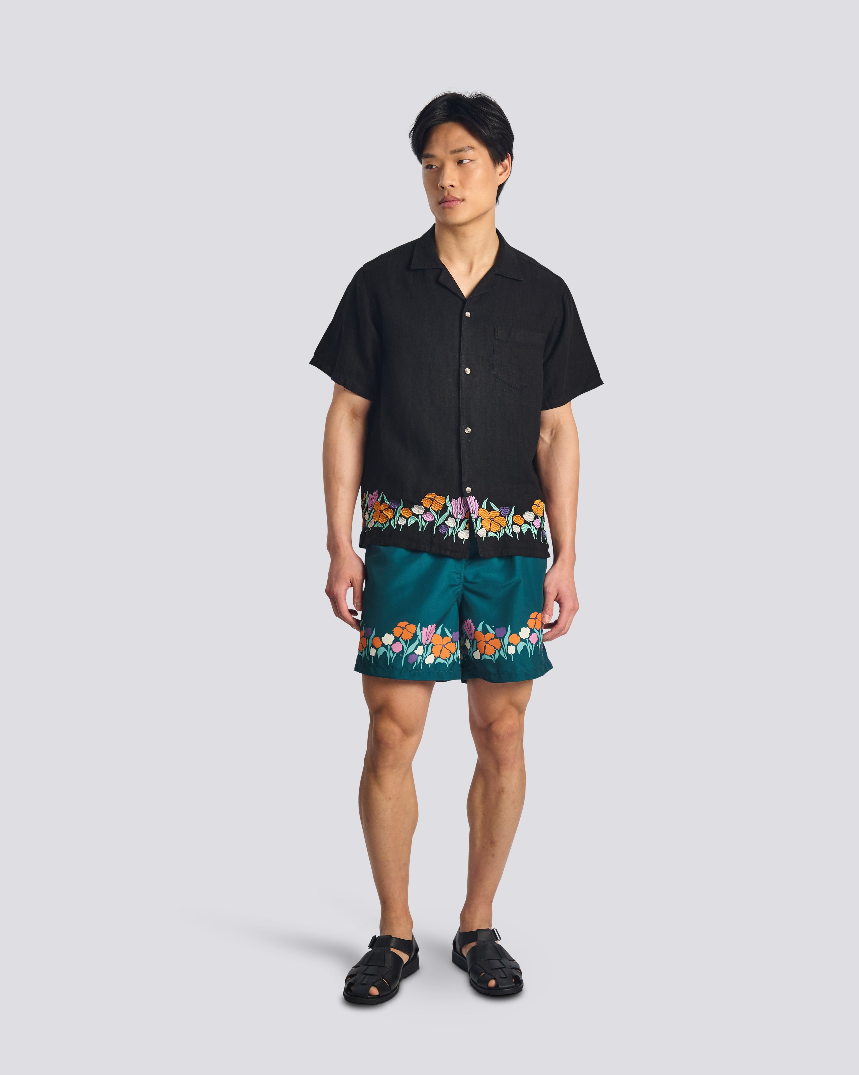 A model wearing the Bather Men's Black Lawn Linen Camp Shirt, styled with matching green floral swim trunks and black leather sandals. The image showcases the shirt's relaxed fit and the vibrant floral embroidery wrapping around the hem.