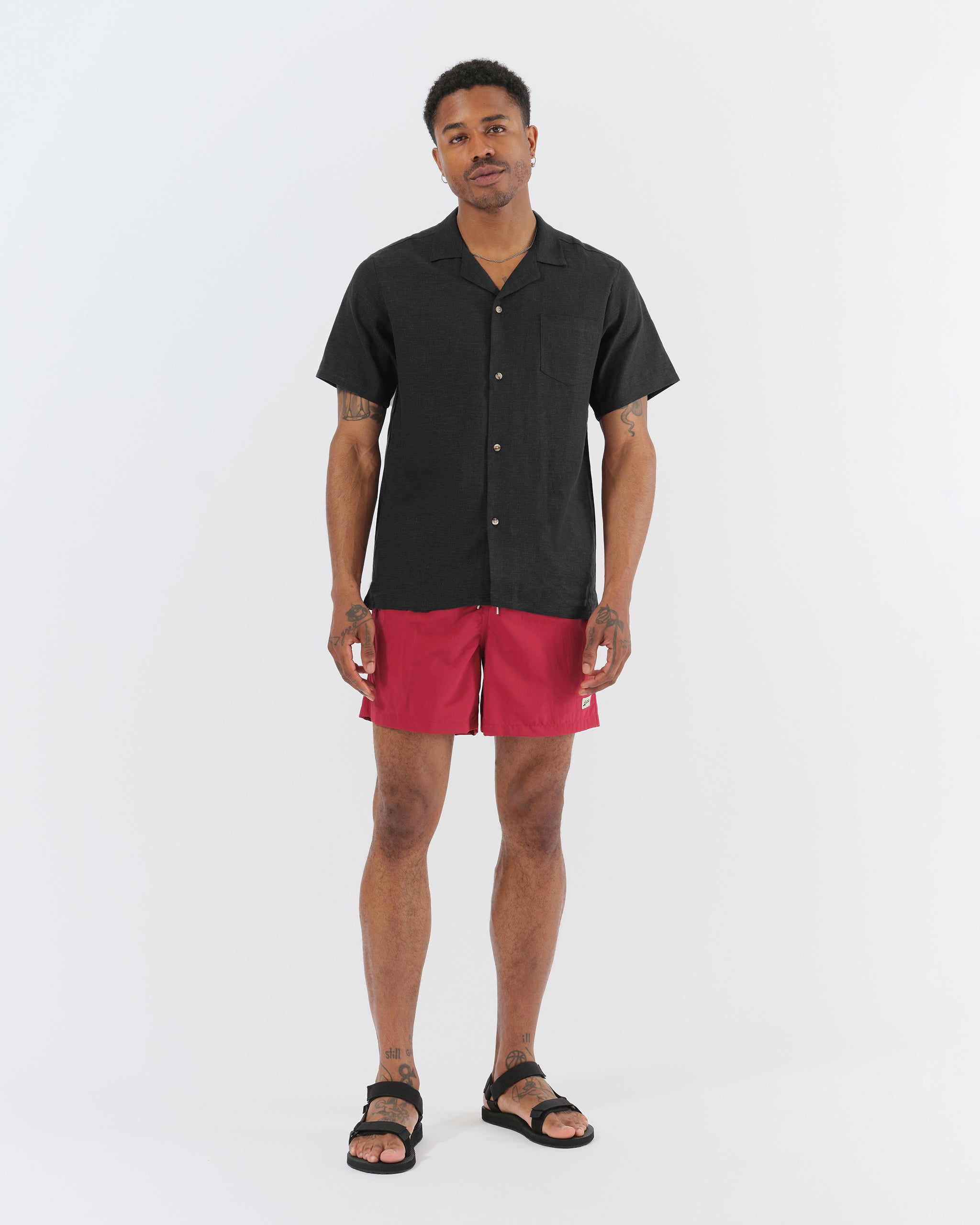 A model wearing the Bather Men's Black Linen Camp Shirt, styled with dark red swim trunks and black leather sandals. The image highlights the shirt's relaxed fit and the natural texture of the 100% black linen fabric.