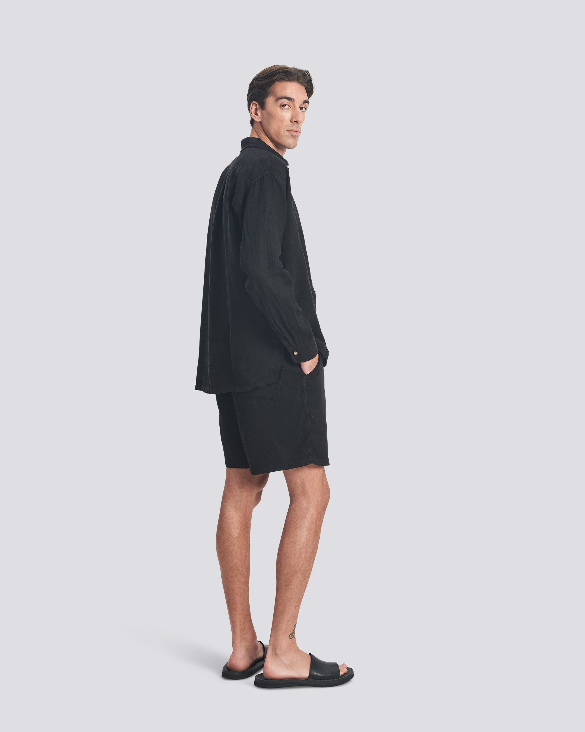 A side-profile view of a model wearing the Bather Men's Black Linen Leisure Shirt, styled with black shorts and black sandals. The image highlights the shirt's long-sleeve silhouette and the natural texture of the midweight 100% black linen fabric.