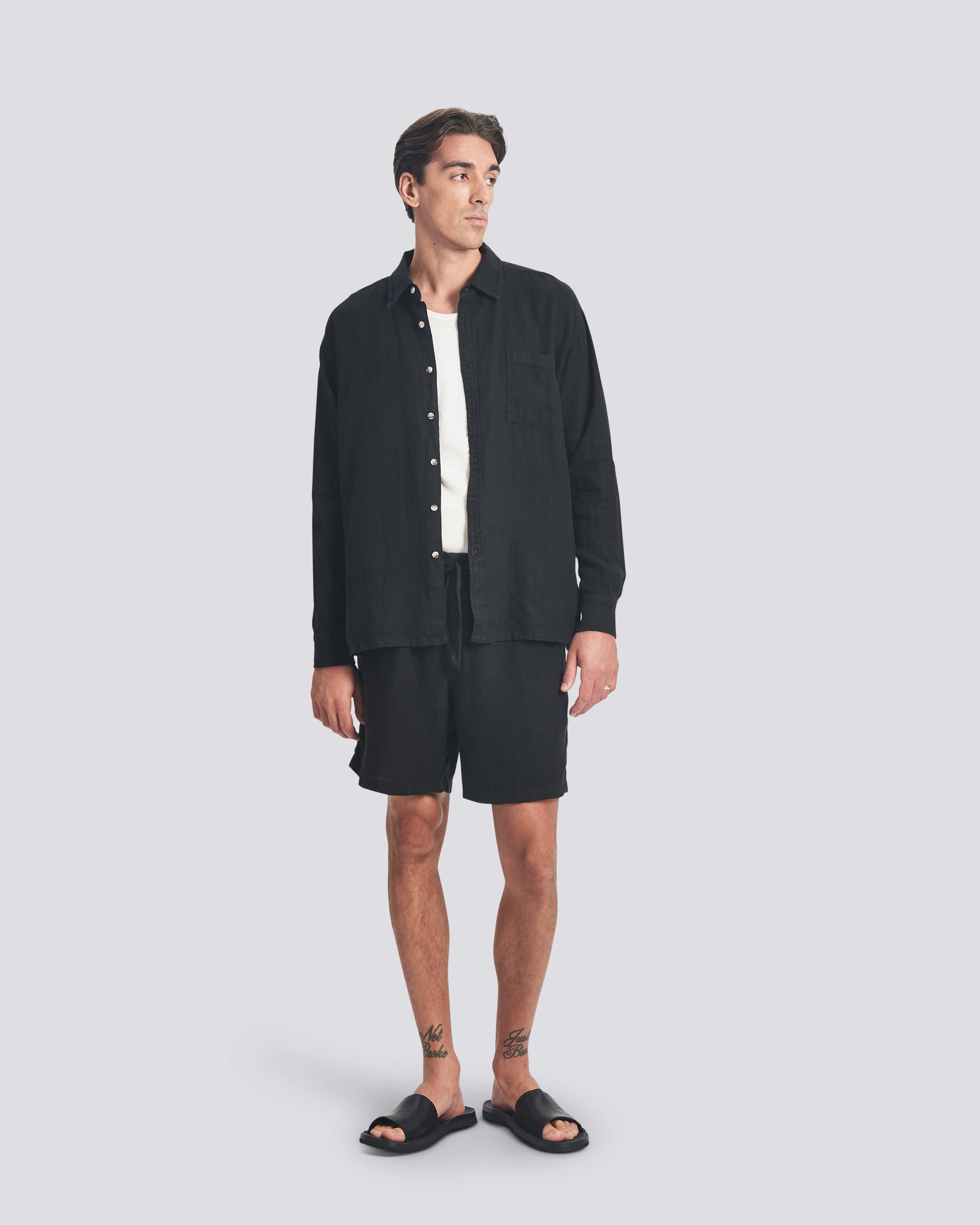 A model wearing the Bather Men's Black Linen Leisure Shirt, layered over a white t-shirt and styled with black shorts and black sandals. The image highlights the shirt's relaxed fit and the natural texture of the midweight 100% black linen fabric.