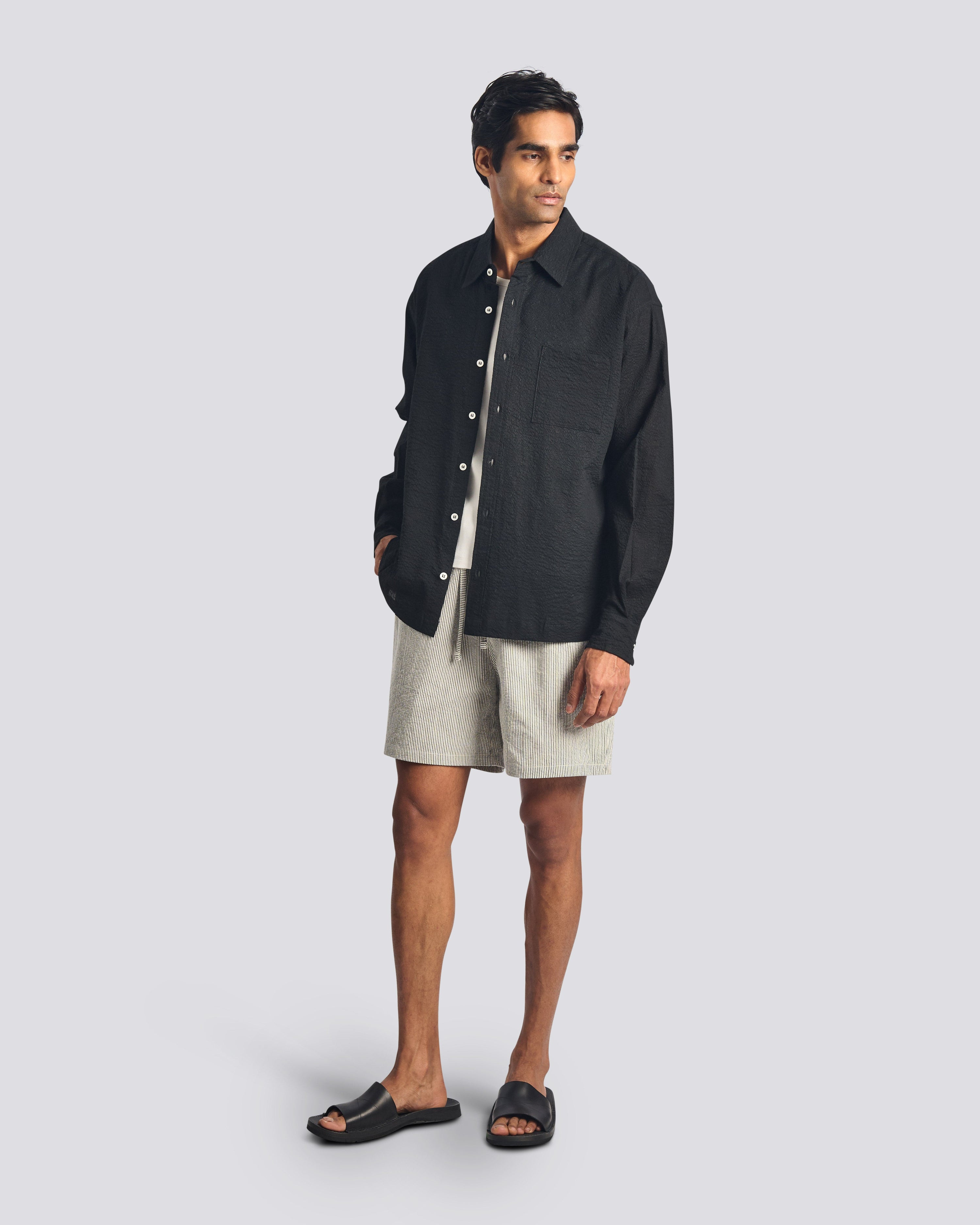A model wearing the Bather Men's Black Ripple Leisure Shirt, layered over a white t-shirt and styled with patterned shorts and black sandals. The image highlights the shirt's relaxed fit and the unique Japanese cotton fabric with a subtle seersucker texture.
