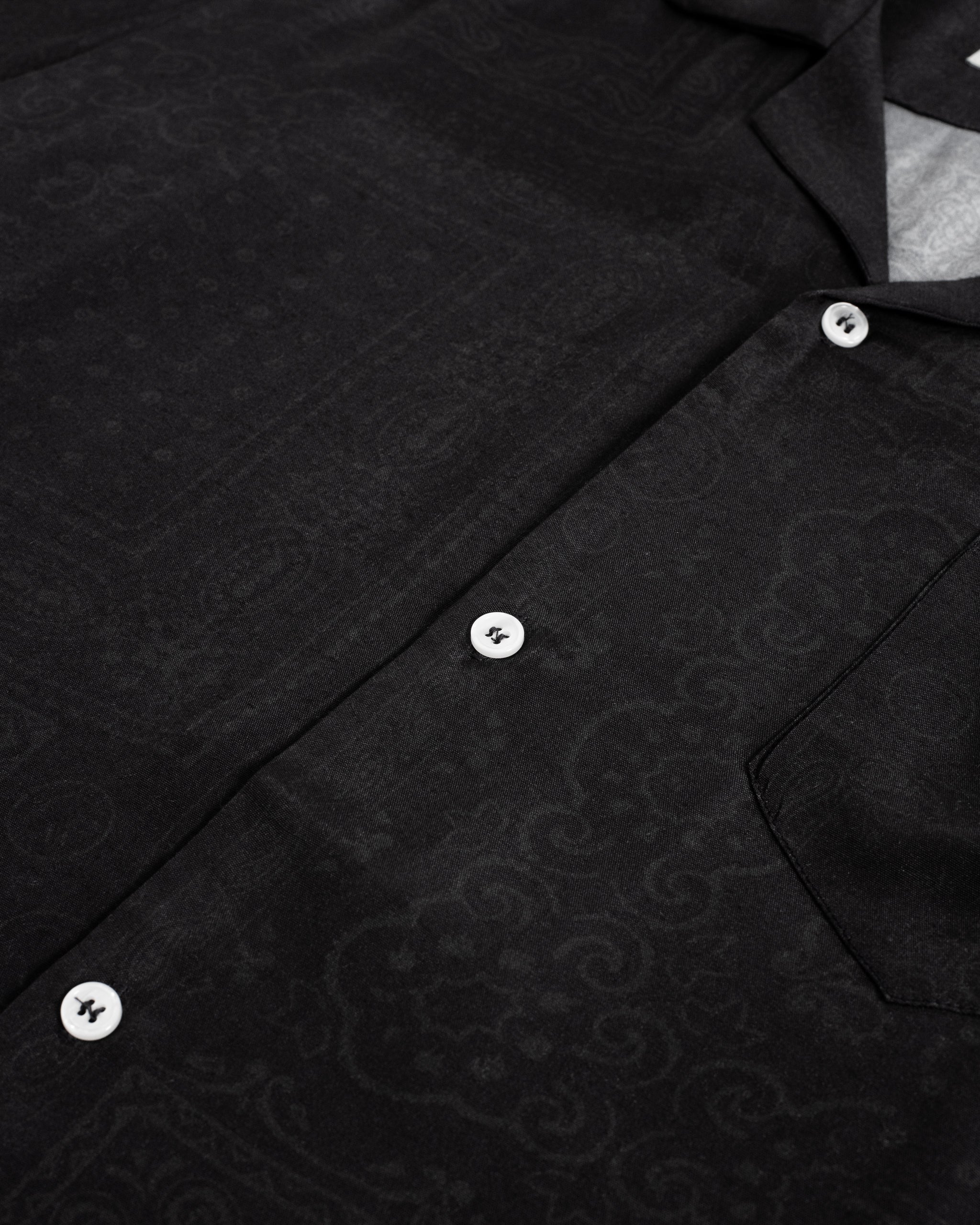 A close-up detail of the Bather Men's Black Tonal Bandana Camp Shirt, showcasing the intricate tonal bandana print on the silky black rayon fabric and the contrast of the white buttons.