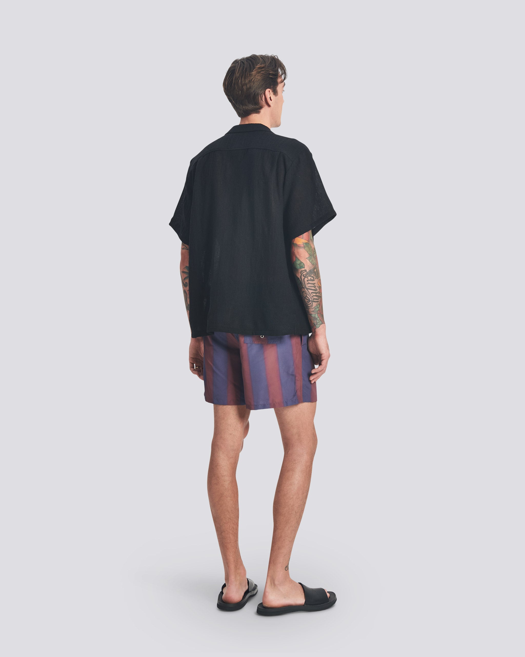Back view of a model wearing the Bather Men's Black Wool Gauze Camp Shirt, highlighting the boxy update and shorter cut of the silhouette. The shirt is styled with purple and brown patterned swim trunks and black sandals.