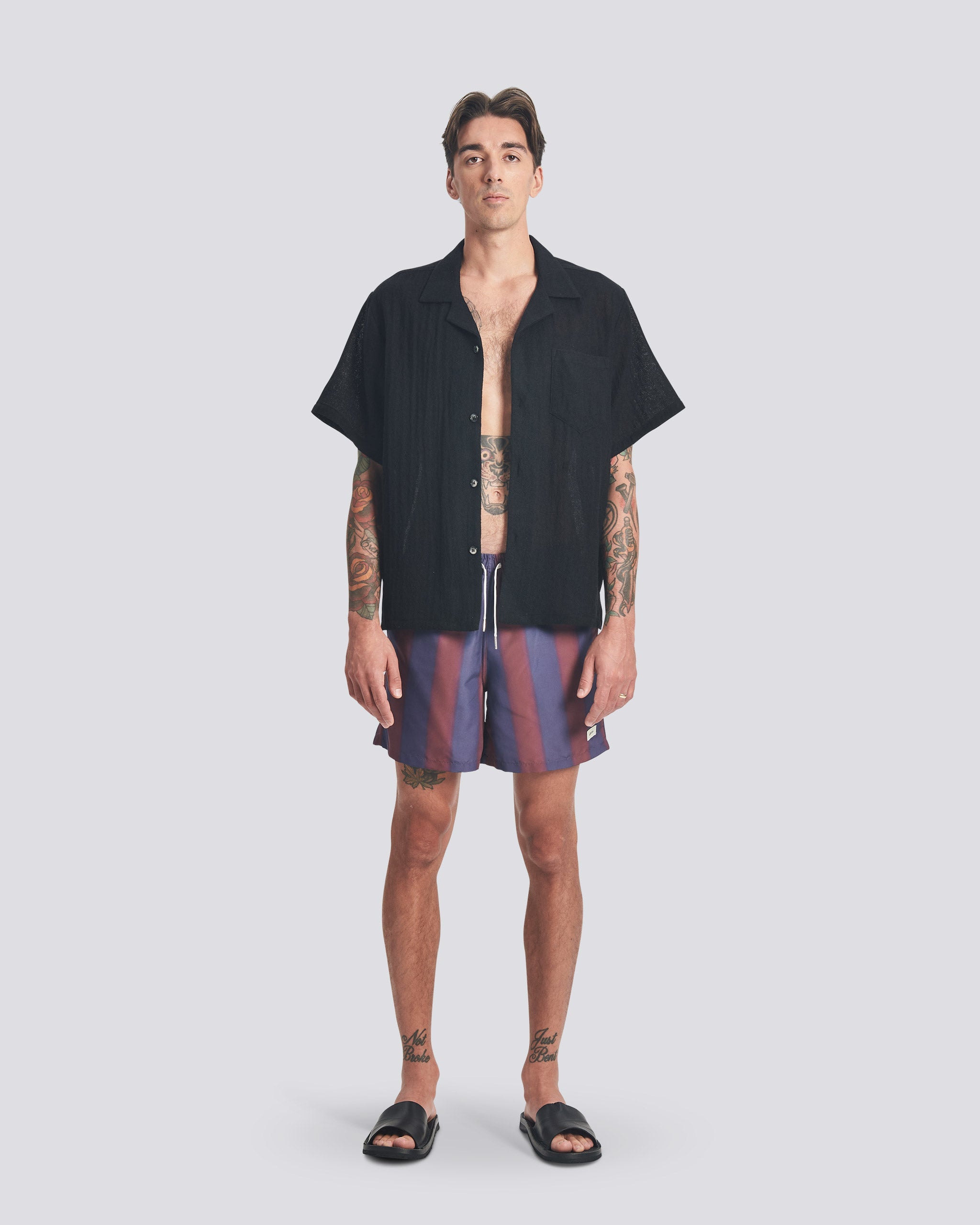 A model wearing the Bather Men's Black Wool Gauze Camp Shirt, styled with purple and brown patterned swim trunks and black sandals. The image highlights the shirt's boxy, relaxed fit and the lightweight texture of the Japanese worsted wool gauze.