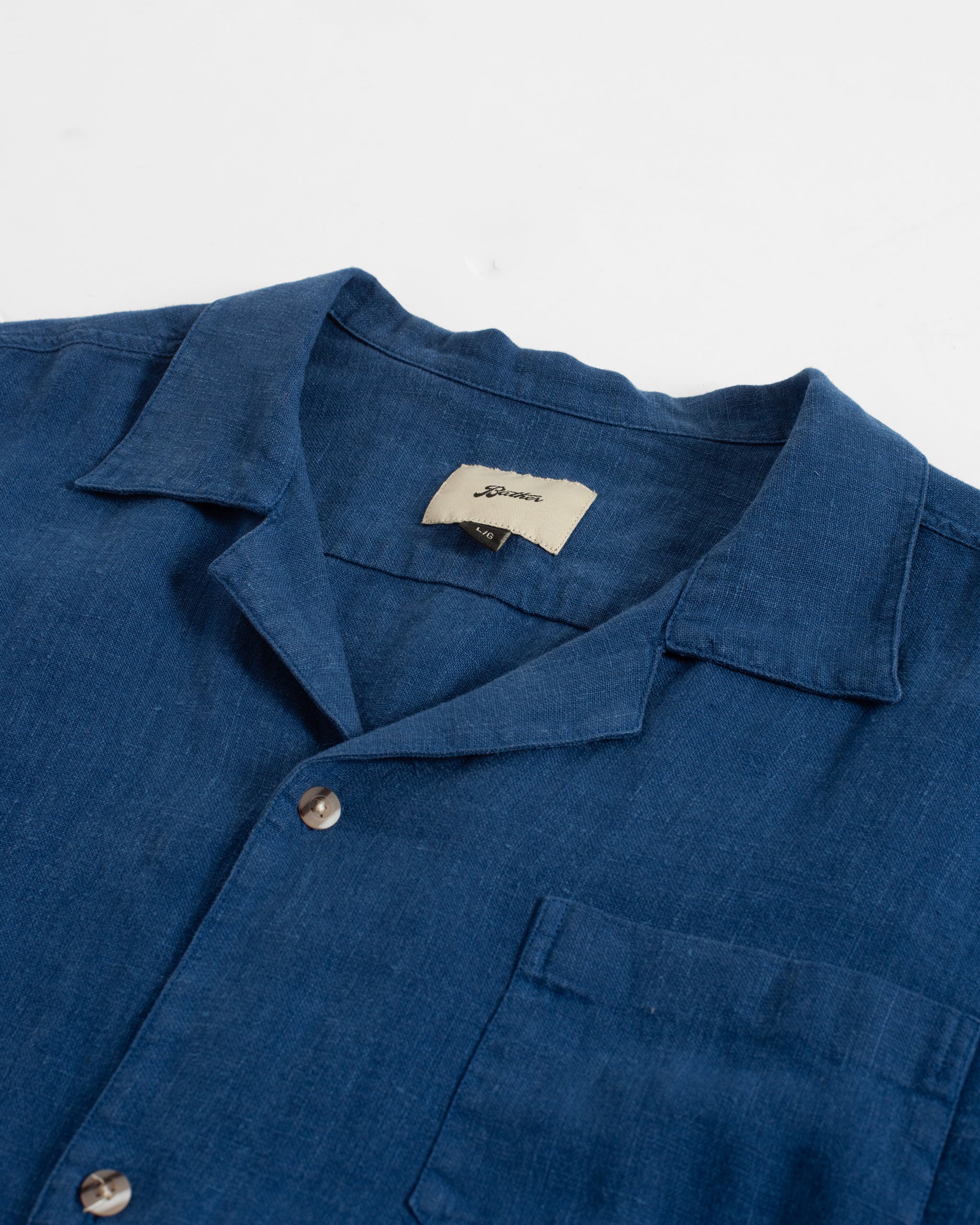 A close-up detail of the Bather Men's Brine Linen Camp Shirt, showcasing the natural texture of the brine blue linen fabric, the classic camp collar with a woven brand label, and the subtle marbled texture of the buttons.