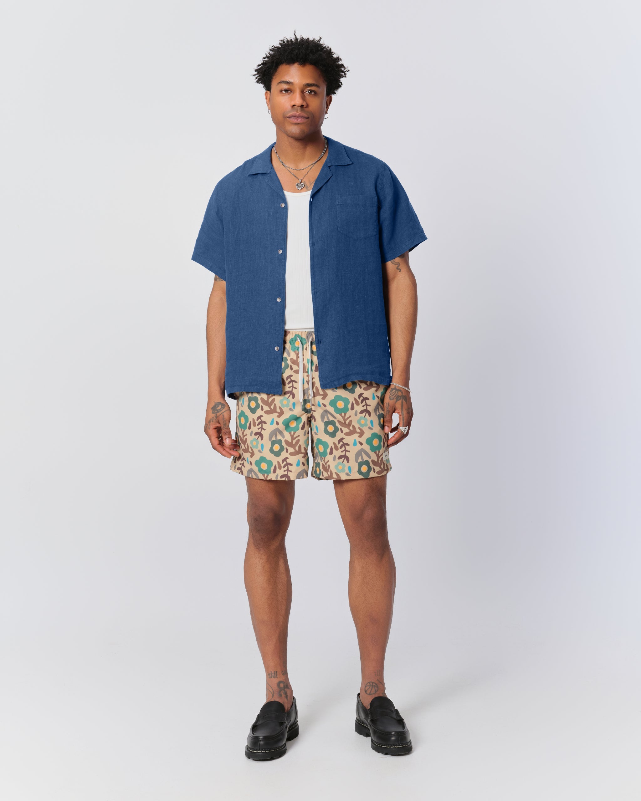 A model wearing the Bather Men's Brine Linen Camp Shirt, layered over a white ribbed tank top and styled with floral patterned shorts and black leather loafers. The image highlights the shirt's relaxed fit and the natural texture of the 100% brine blue linen fabric.