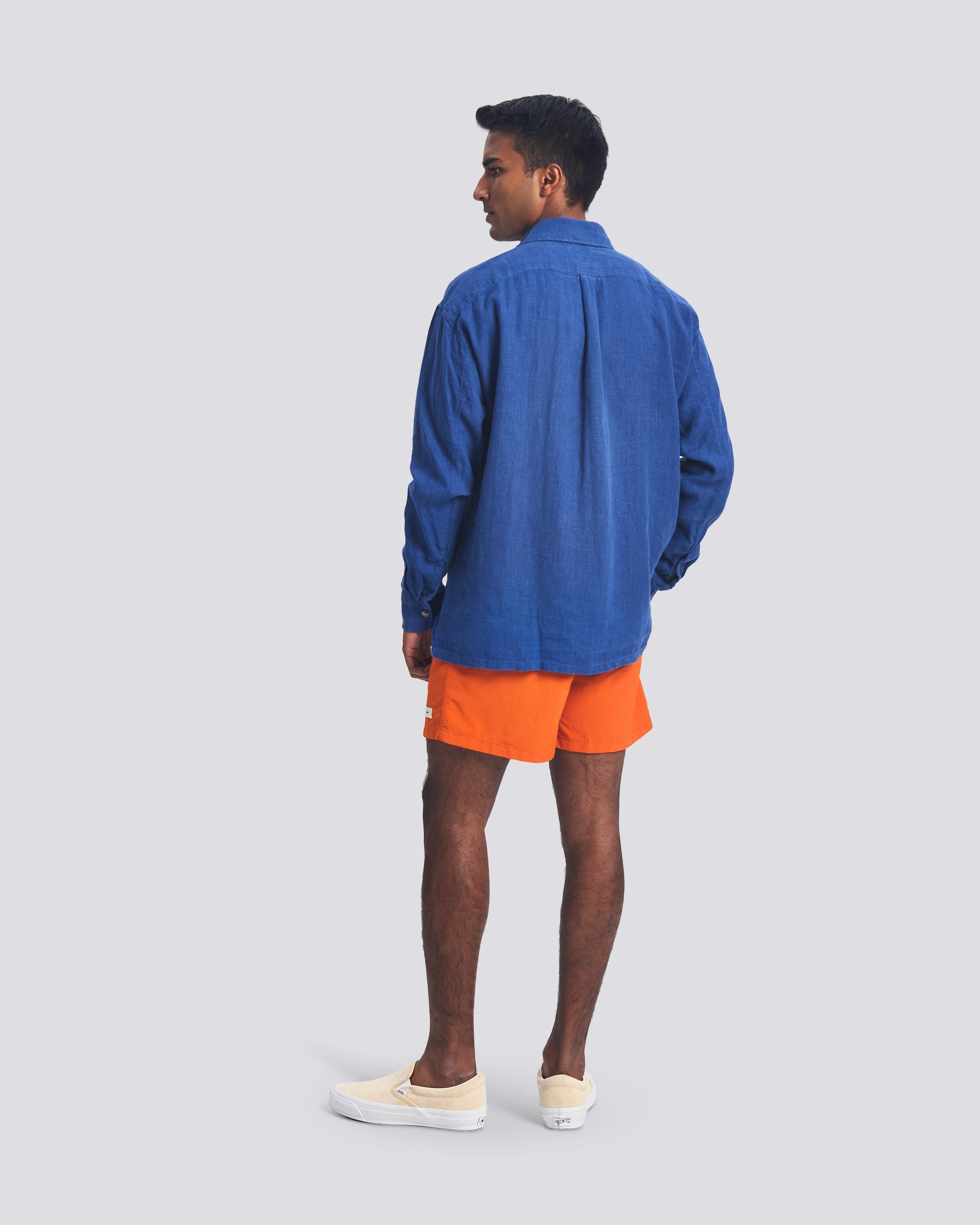 Back view of a model wearing the Bather Men's Brine Linen Leisure Shirt, highlighting the relaxed fit, long sleeves, and straight hem. The shirt is styled with bright orange shorts and cream slip-on sneakers against a white background.