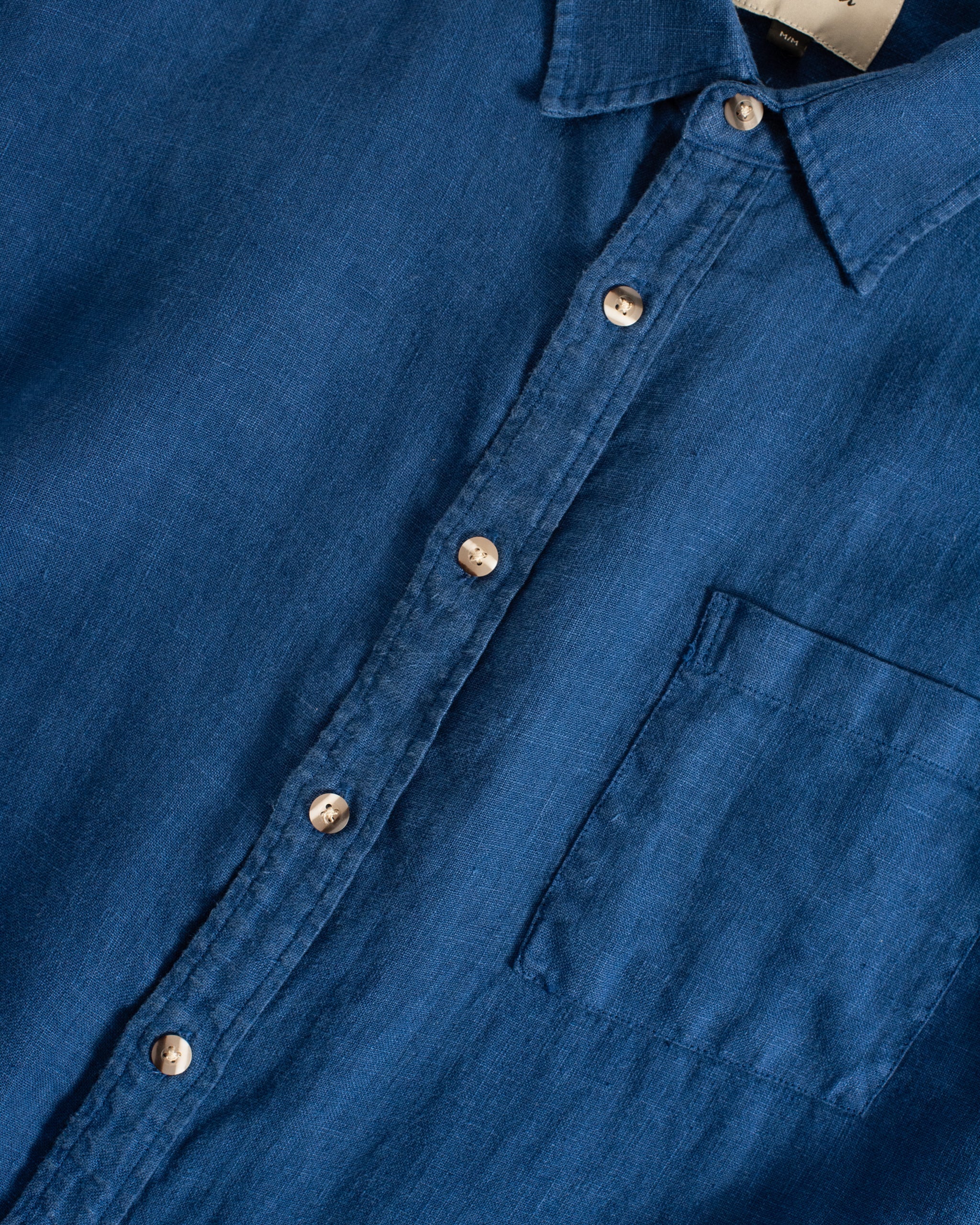 A close-up detail of the Bather Men's Brine Linen Leisure Shirt, showcasing the natural texture of the midweight brine blue linen fabric, the classic collar, and the contrast of the marbled light-colored buttons.