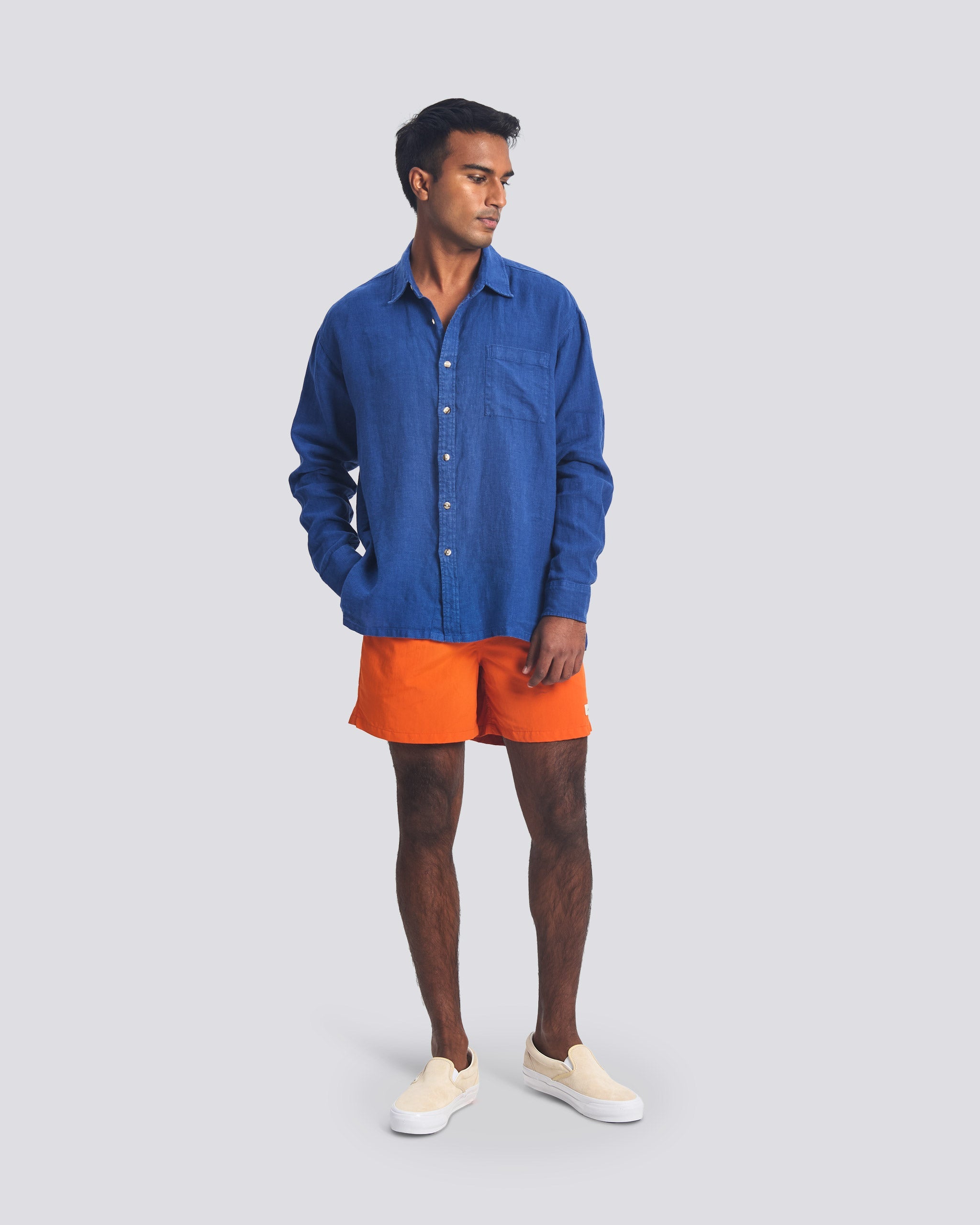 A model wearing the Bather Men's Brine Linen Leisure Shirt, styled with bright orange shorts and cream slip-on sneakers. The image highlights the shirt's relaxed fit, long-sleeve silhouette, and the natural texture of the midweight 100% brine blue linen fabric.