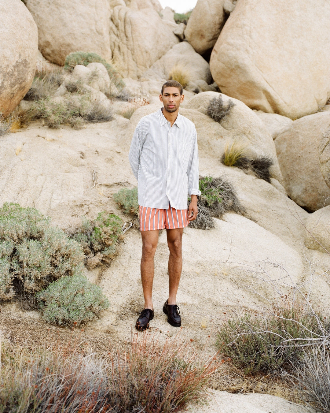 Male model wearing light blue and white striped button-up shirt and red striped swim trunks standing on rocky desert terrain