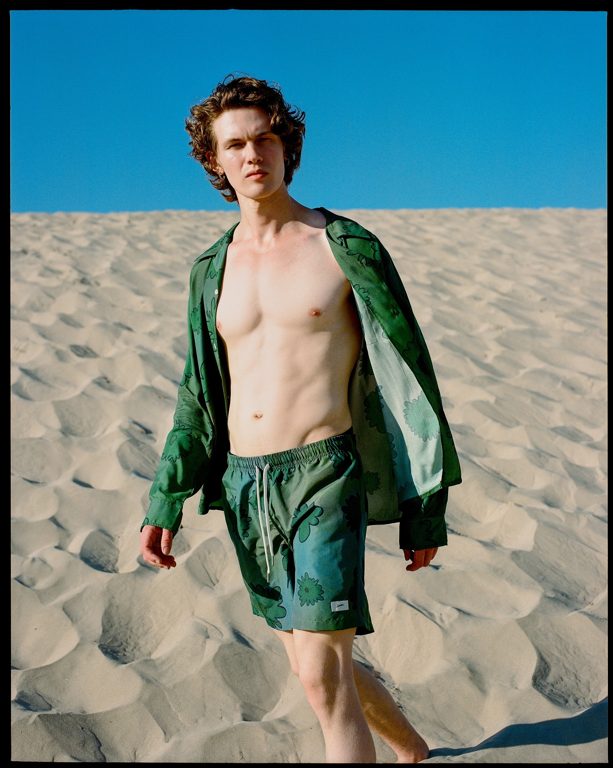 Model wearing an unbuttoned Bather Men's green floral long-sleeve camp shirt and matching shorts in a desert-like beach setting.