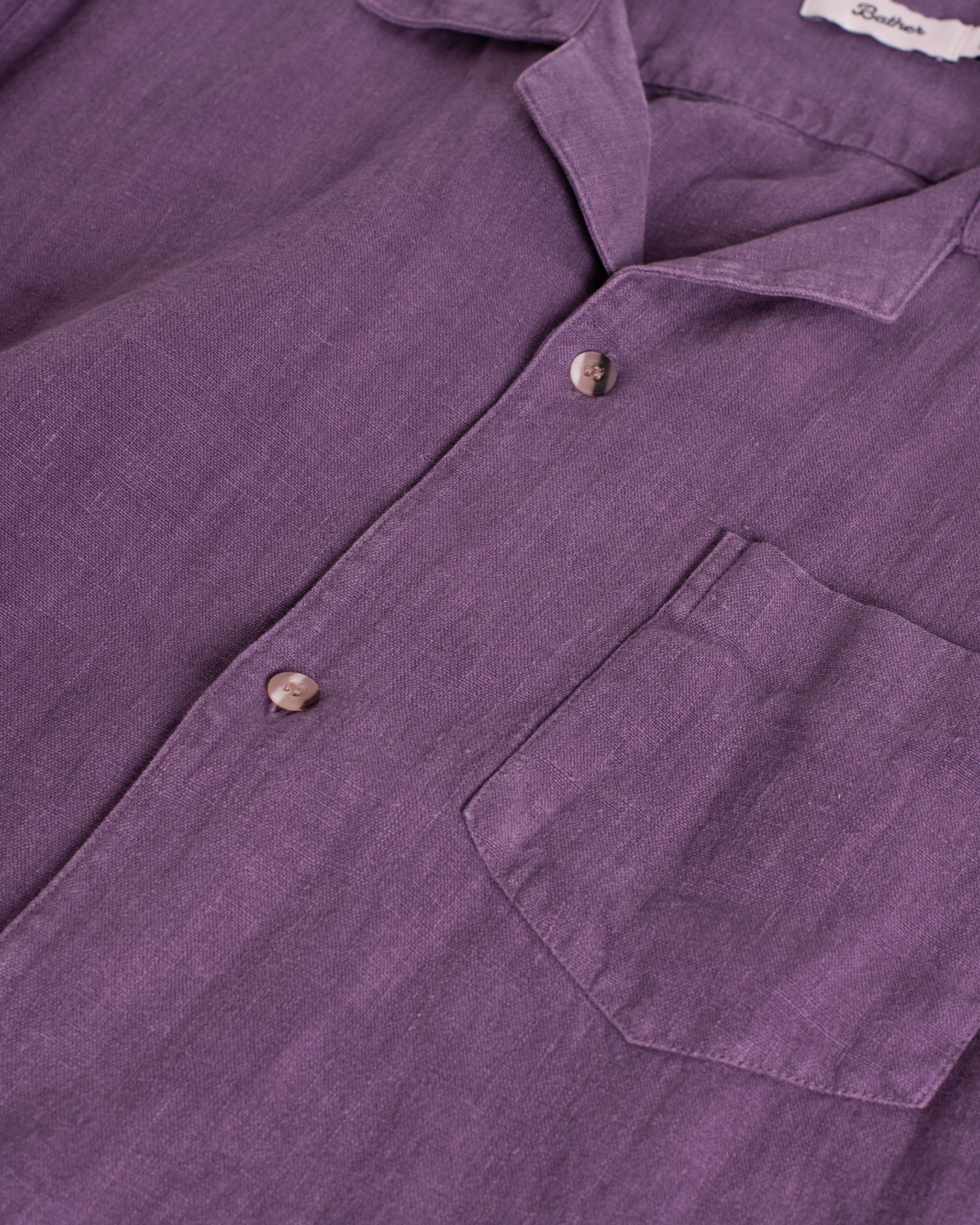 Close-up of the Bather Men's Grape Linen Camp Shirt in muted purple, showing the textured linen fabric, a chest pocket, tortoise-shell buttons, and the Bather neck label.
