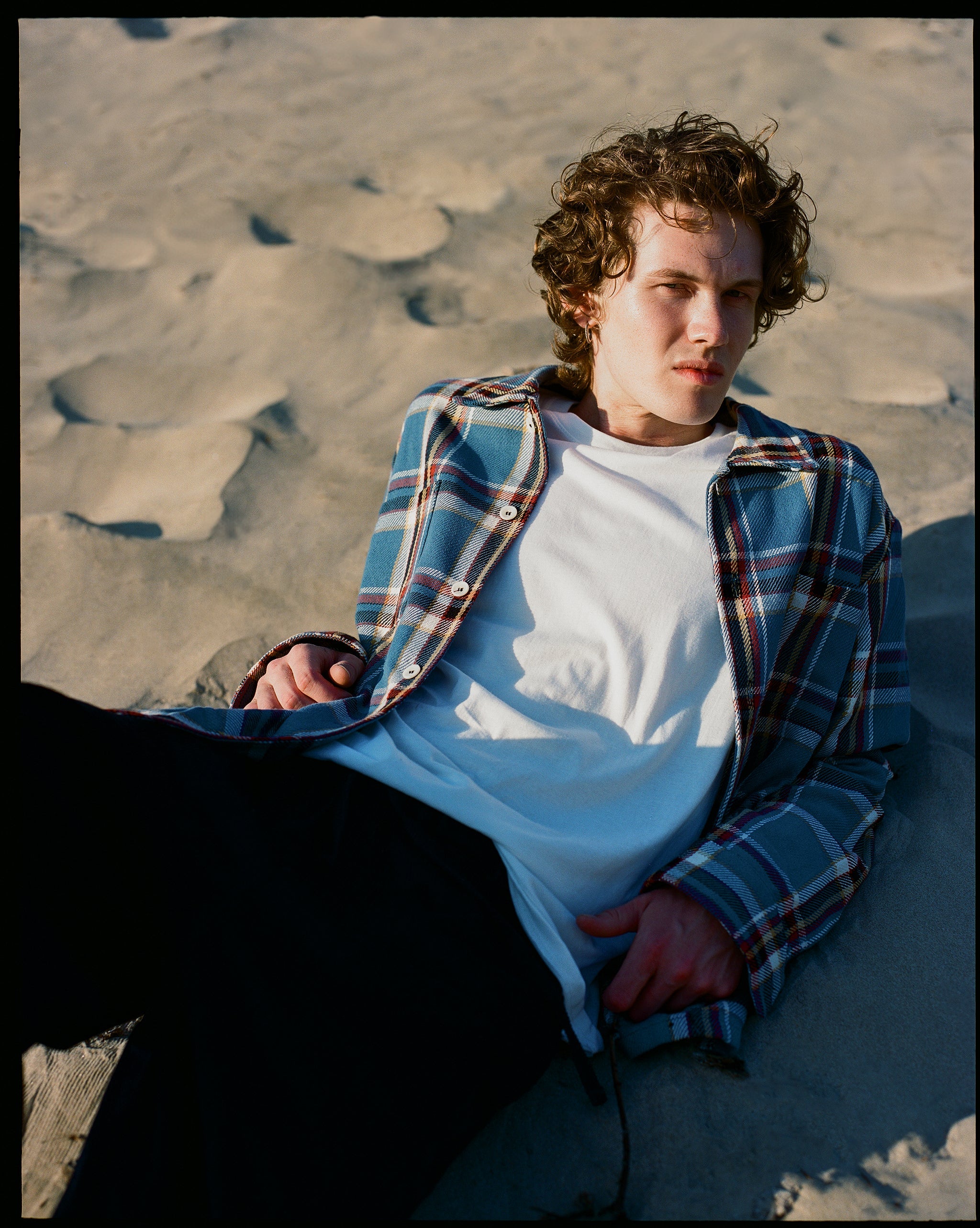 Bather Men's Marlin Plaid Flannel Shirt in a blue and tan checkered pattern, worn open over a white t-shirt by a model sitting on a sandy beach.