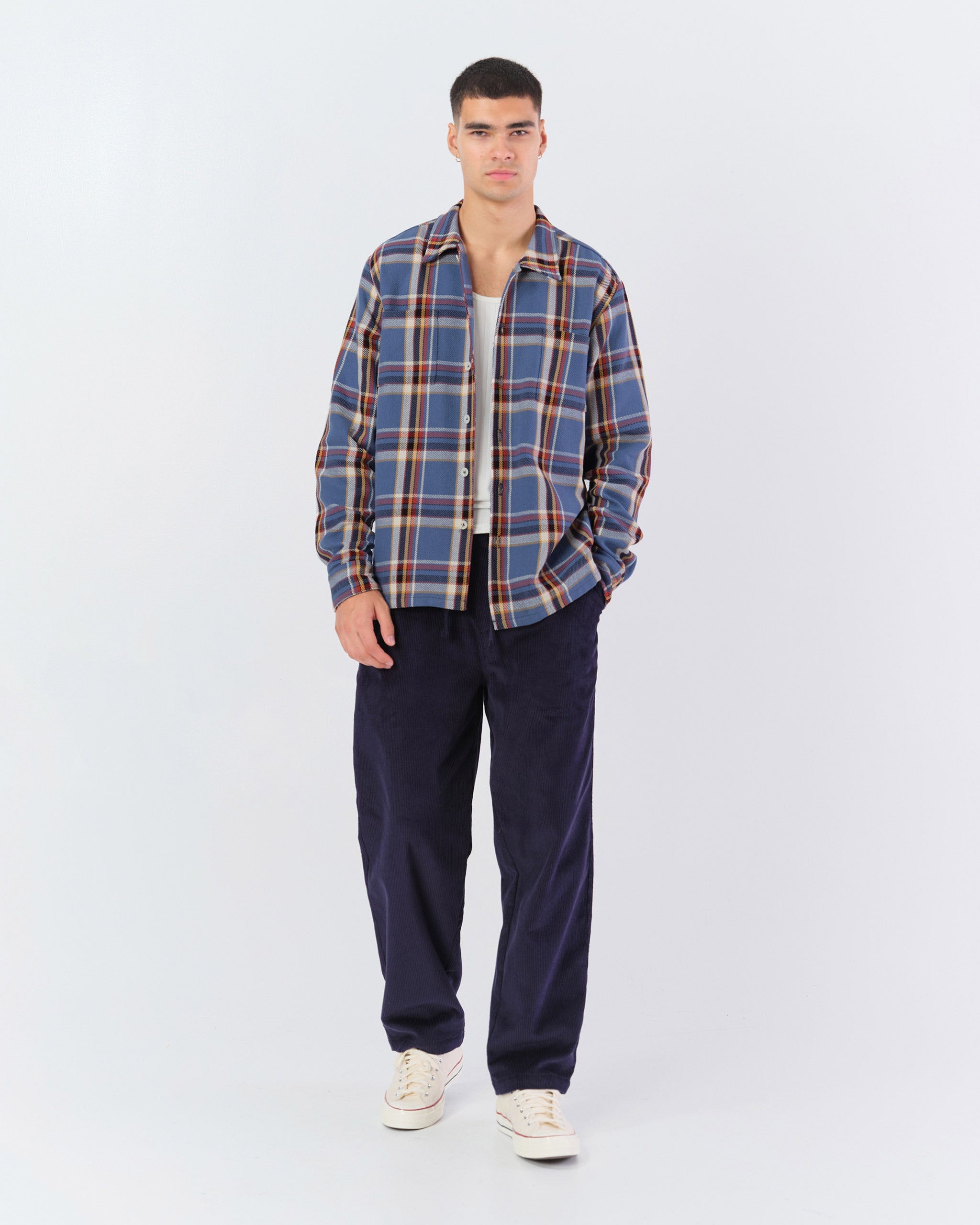 Model in Bather Men's Marlin Plaid Flannel Shirt in a blue and tan checkered pattern, styled open over a white tank top with dark navy corduroy trousers and white sneakers on a model.