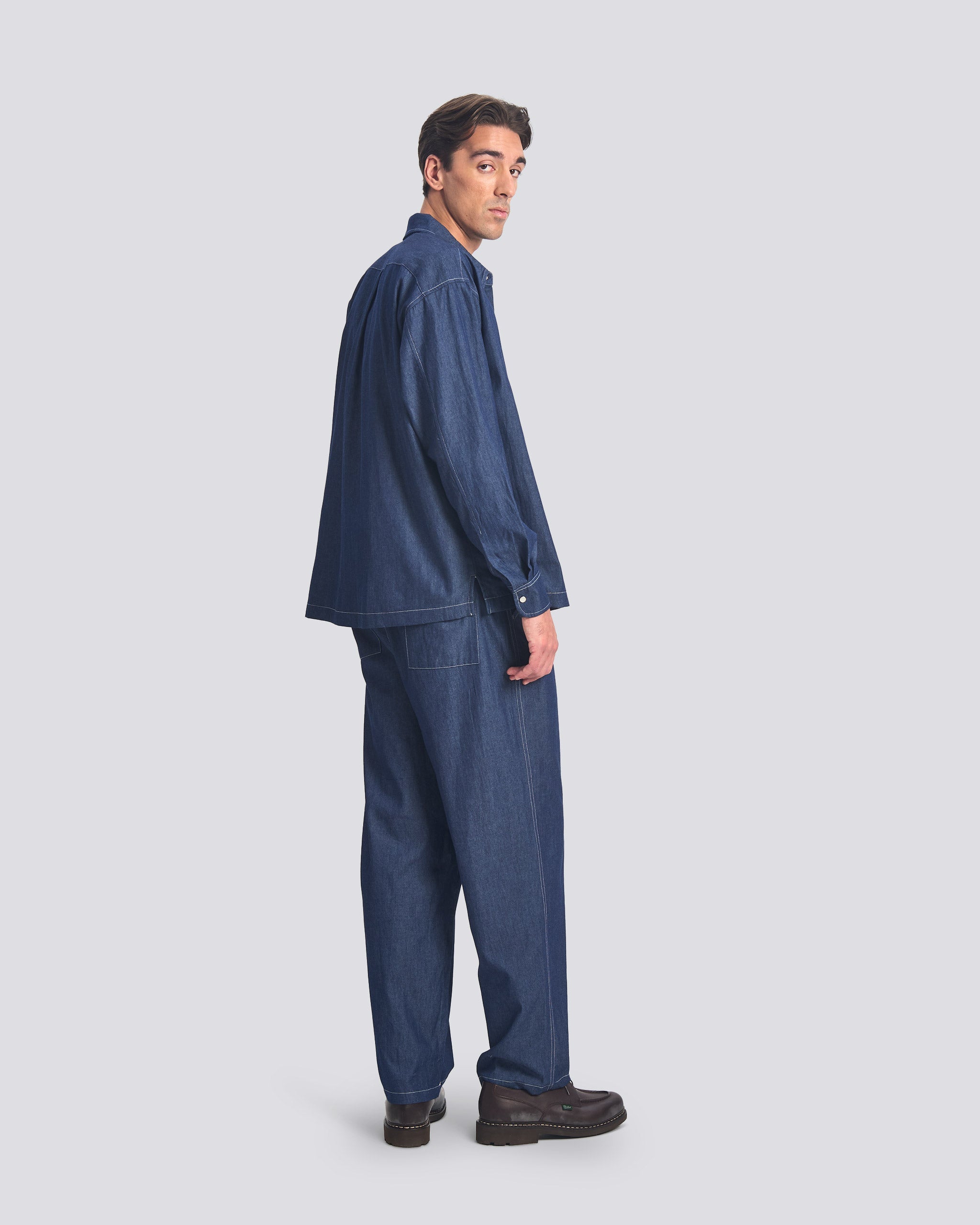 Bather Men's Rinsed Denim Leisure Shirt in dark indigo, showing a rear view of the oversized fit and contrast stitching on a model wearing matching denim pants.