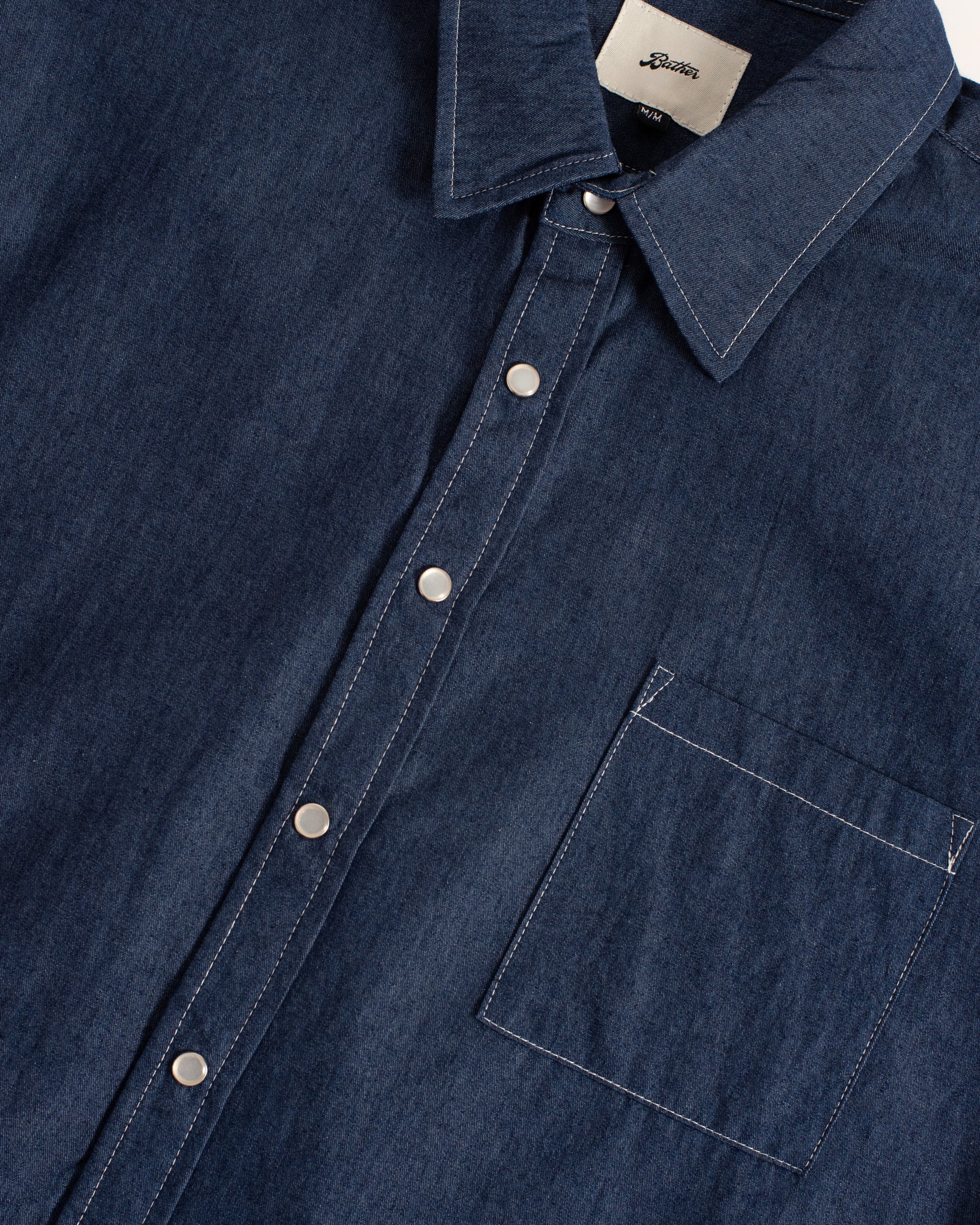 Close-up of the Bather Men's Rinsed Denim Leisure Shirt in dark indigo, showing the fine weave of the 10 oz Japanese denim, white snap buttons, and contrast stitching.