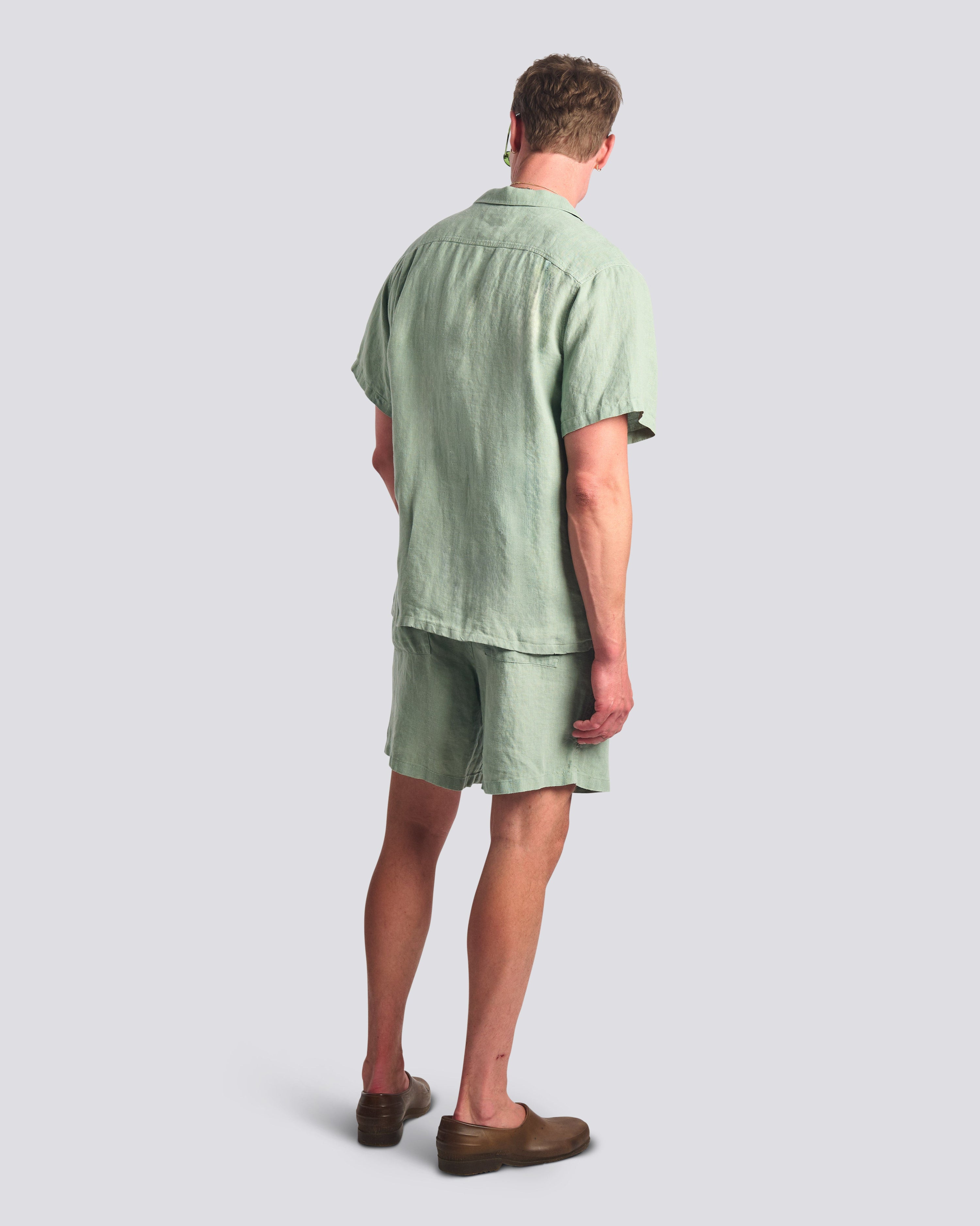 Bather Men's Sage Linen Camp Shirt in light green, showing a rear view of the regular fit and textured linen fabric on a model wearing matching Sage shorts.