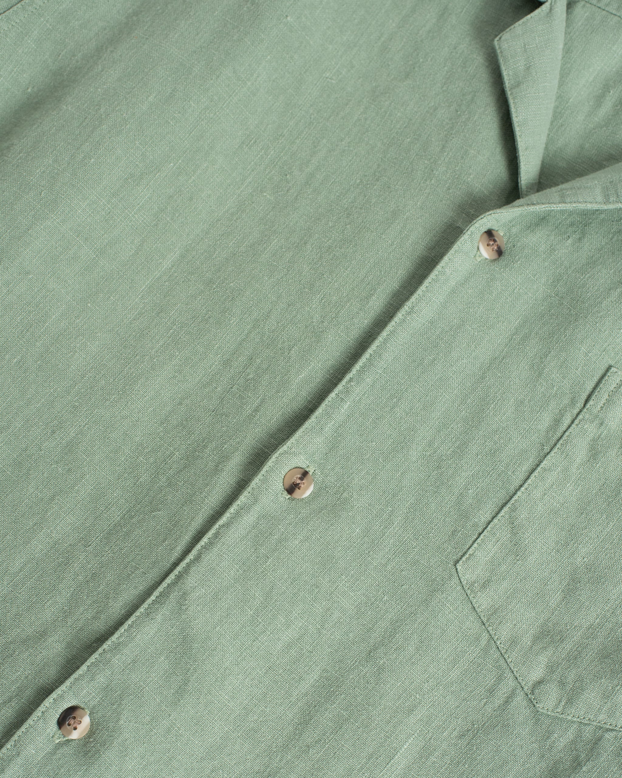 Close-up of the Bather Men's Sage Linen Camp Shirt in light green, highlighting the textured 100% linen weave, brown mottled buttons, and the edge of the front chest pocket.