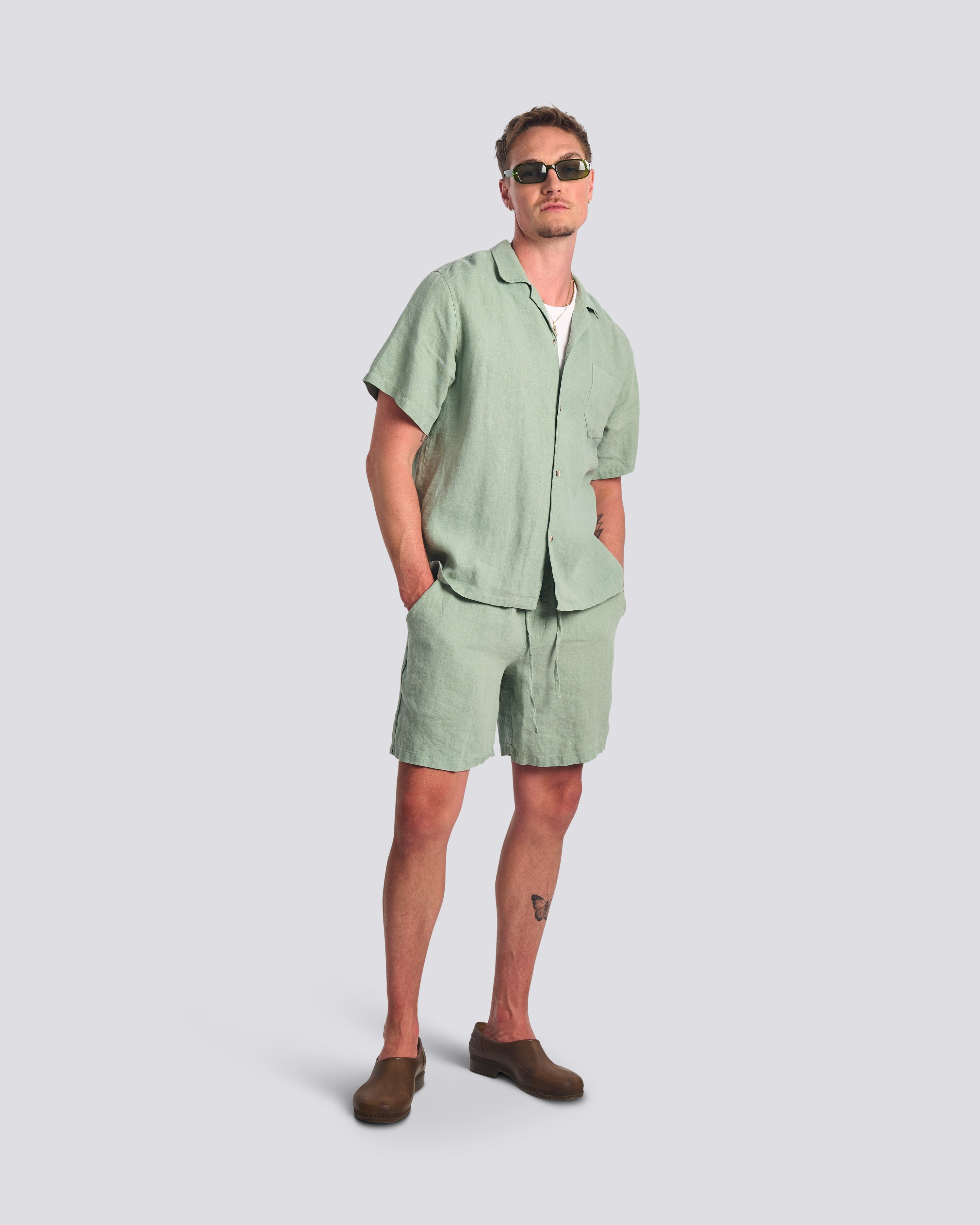 Bather Men's Sage Linen Camp Shirt in light green, styled as a matching set with Sage linen shorts over a white t-shirt and brown loafers on a model wearing sunglasses.