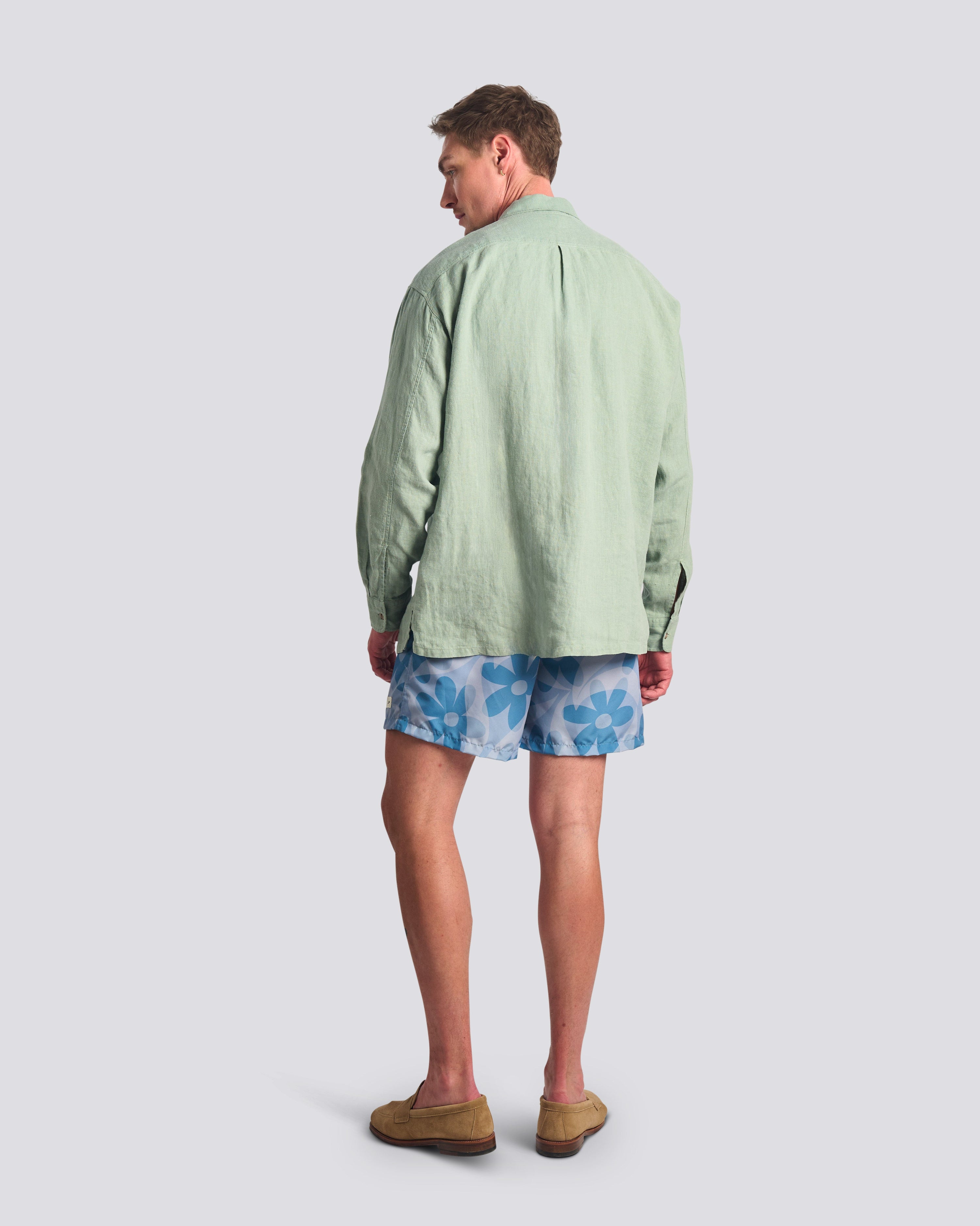 Rear view of the Bather Men's Sage Linen Leisure Shirt in light green, showcasing the regular fit and natural linen texture on a model wearing blue floral swim trunks.