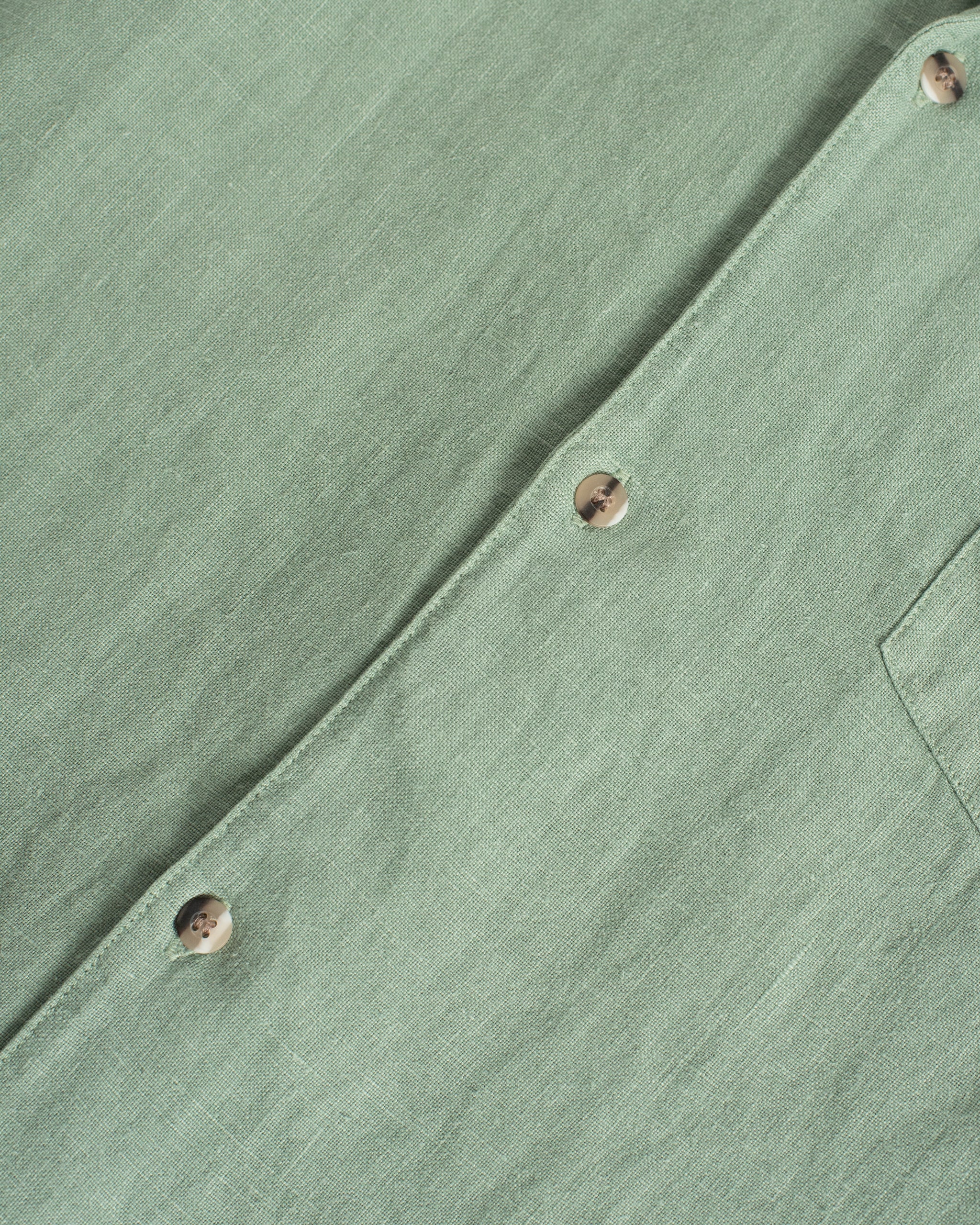 Close-up of the Bather Men's Sage Linen Leisure Shirt in light green, showcasing the textured linen fabric, a front chest pocket, and brown mottled buttons.