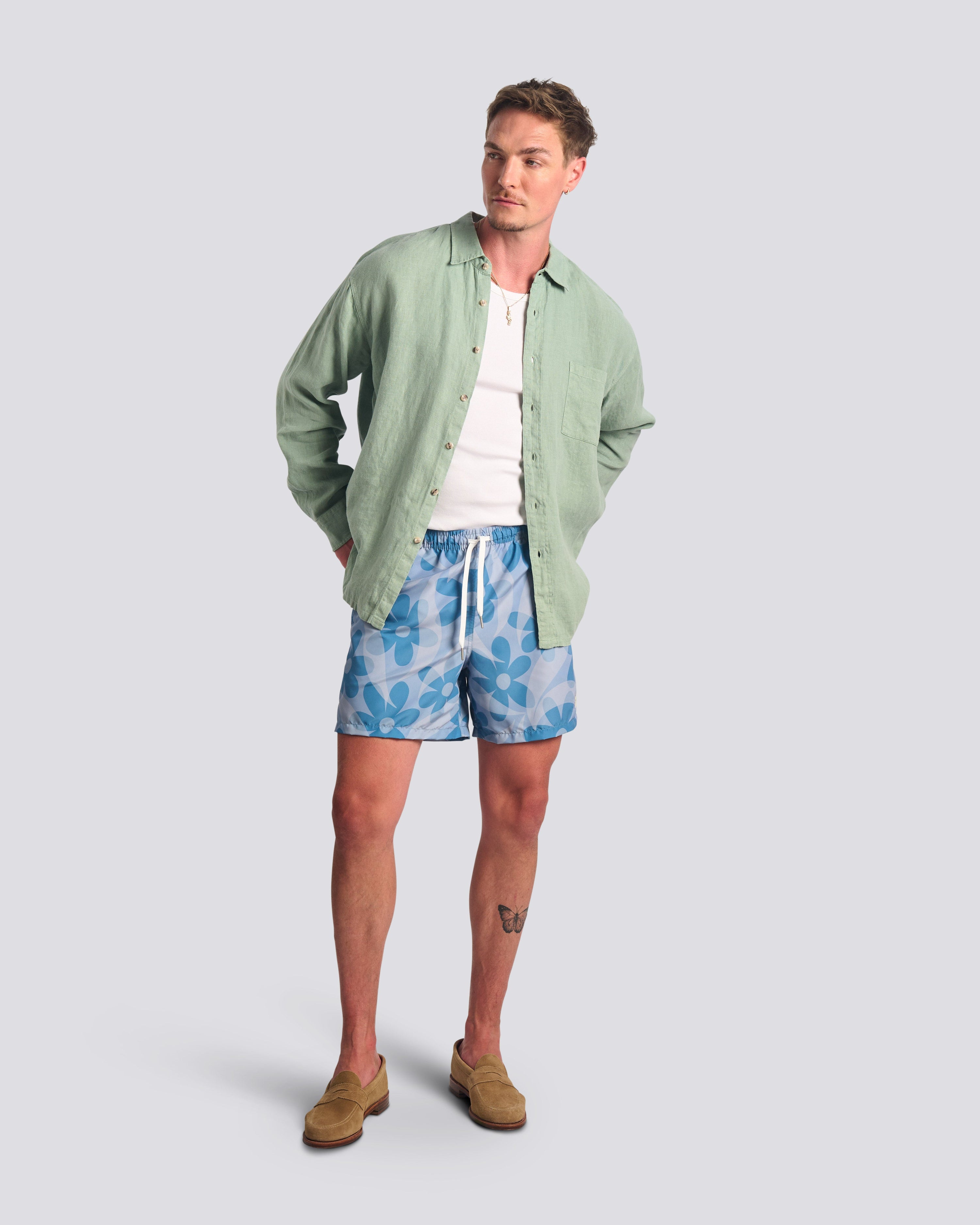 Bather Men's Sage Linen Leisure Shirt in light green, styled open over a white ribbed tank top with blue floral swim trunks and tan loafers on a model.
