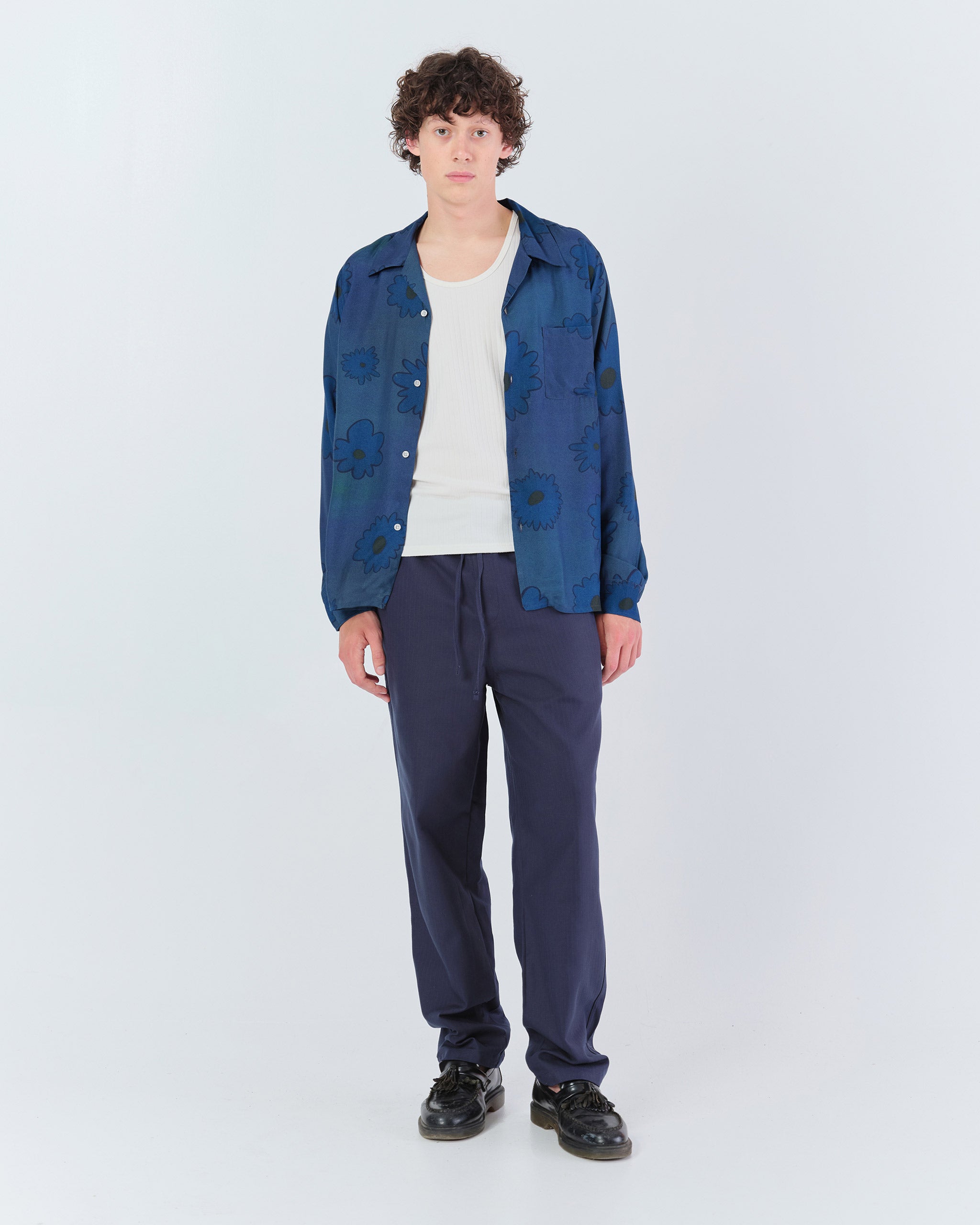 Bather Men’s Sapphire Bloomscape Long Sleeve Camp Shirt in a dark blue floral print, styled open over a white tank top with navy drawstring trousers and black leather shoes on a model.