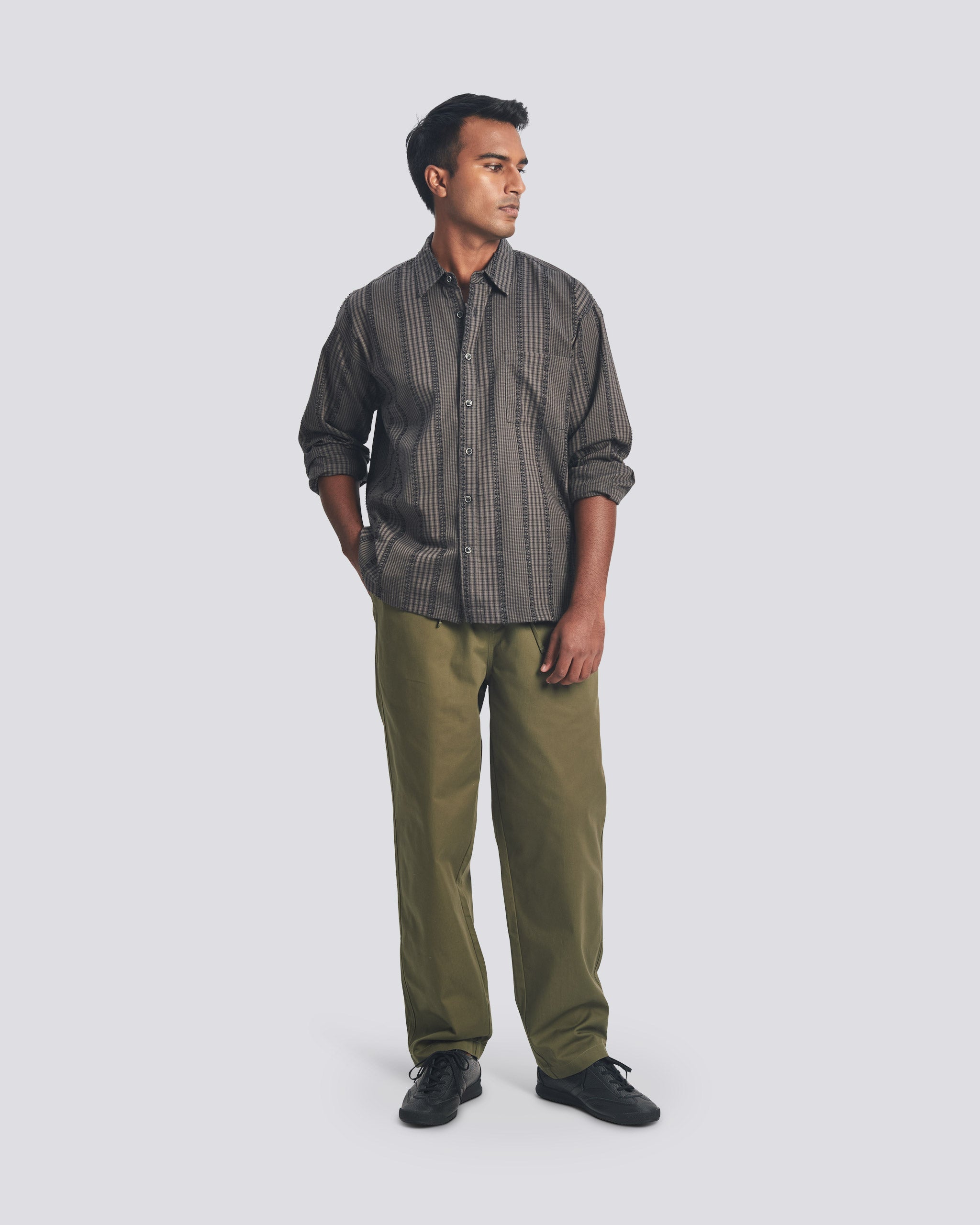 Bather Men’s Smoke Fringe Leisure Shirt in charcoal grey, styled with olive green trousers and black shoes on a model standing against a light grey background.