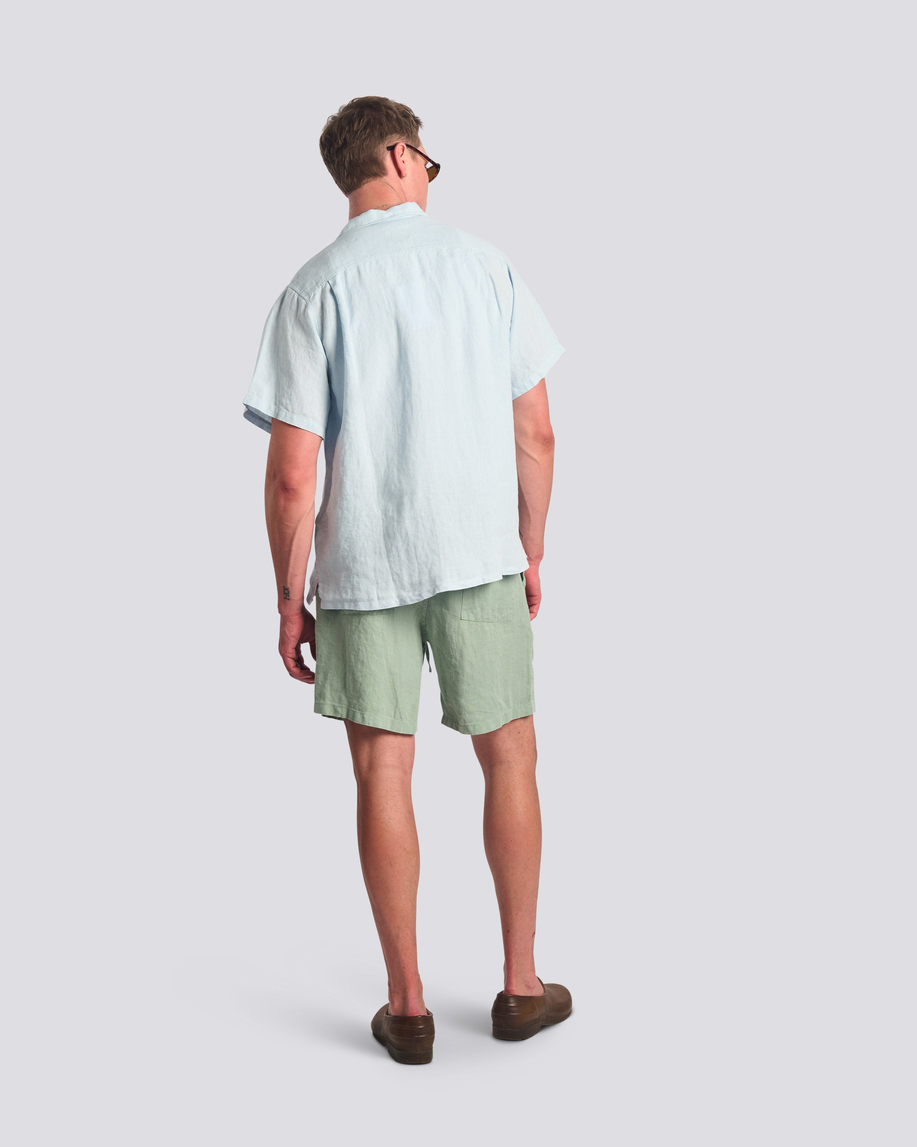 Rear view of the Bather Men's Sky Linen Camp Shirt in light blue, showing the regular fit and textured linen on a model wearing Sage linen shorts and brown loafers.