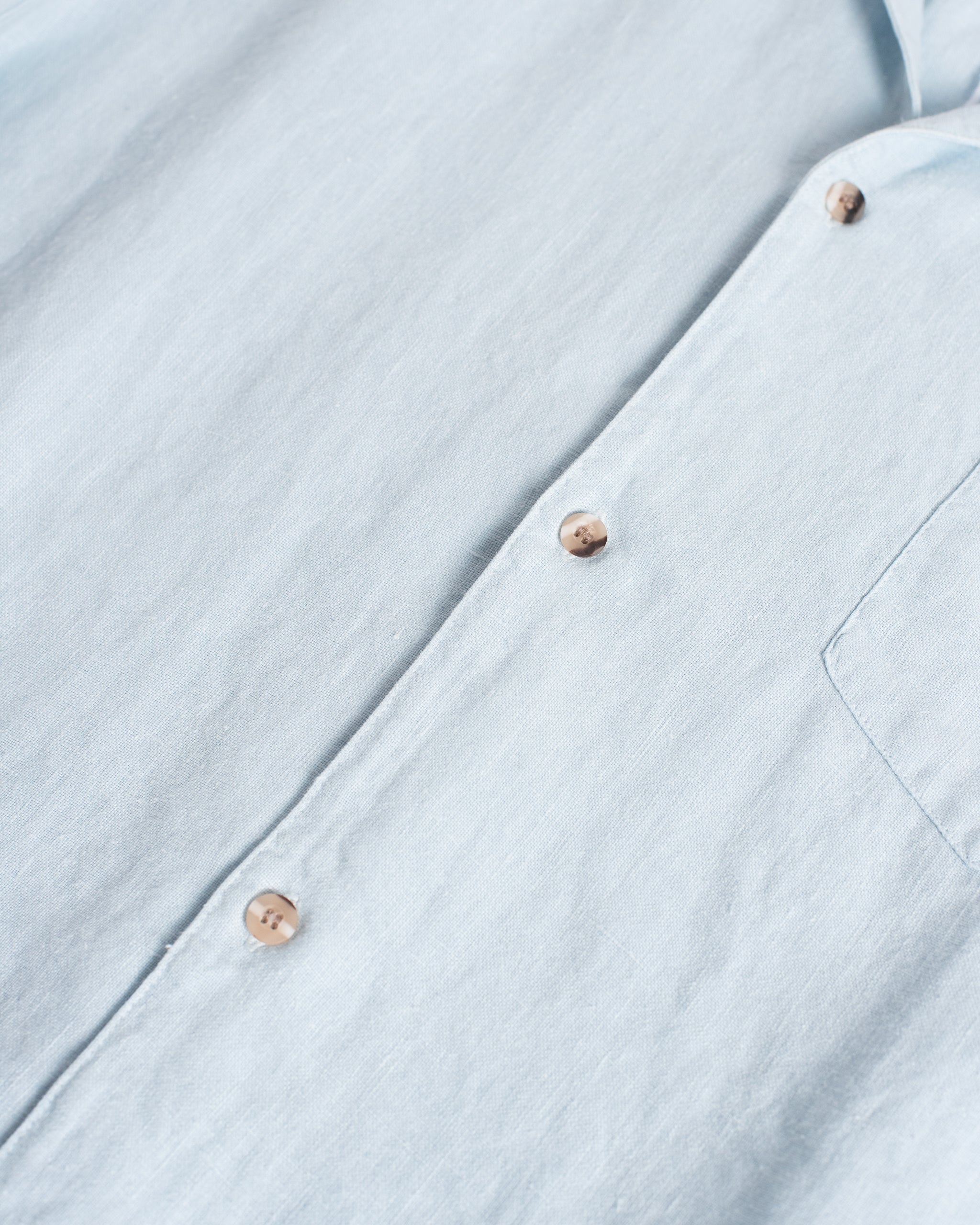 Close-up of the Bather Men's Sky Linen Camp Shirt in light blue, showing the textured 100% linen weave, brown mottled buttons, and the edge of the front chest pocket.