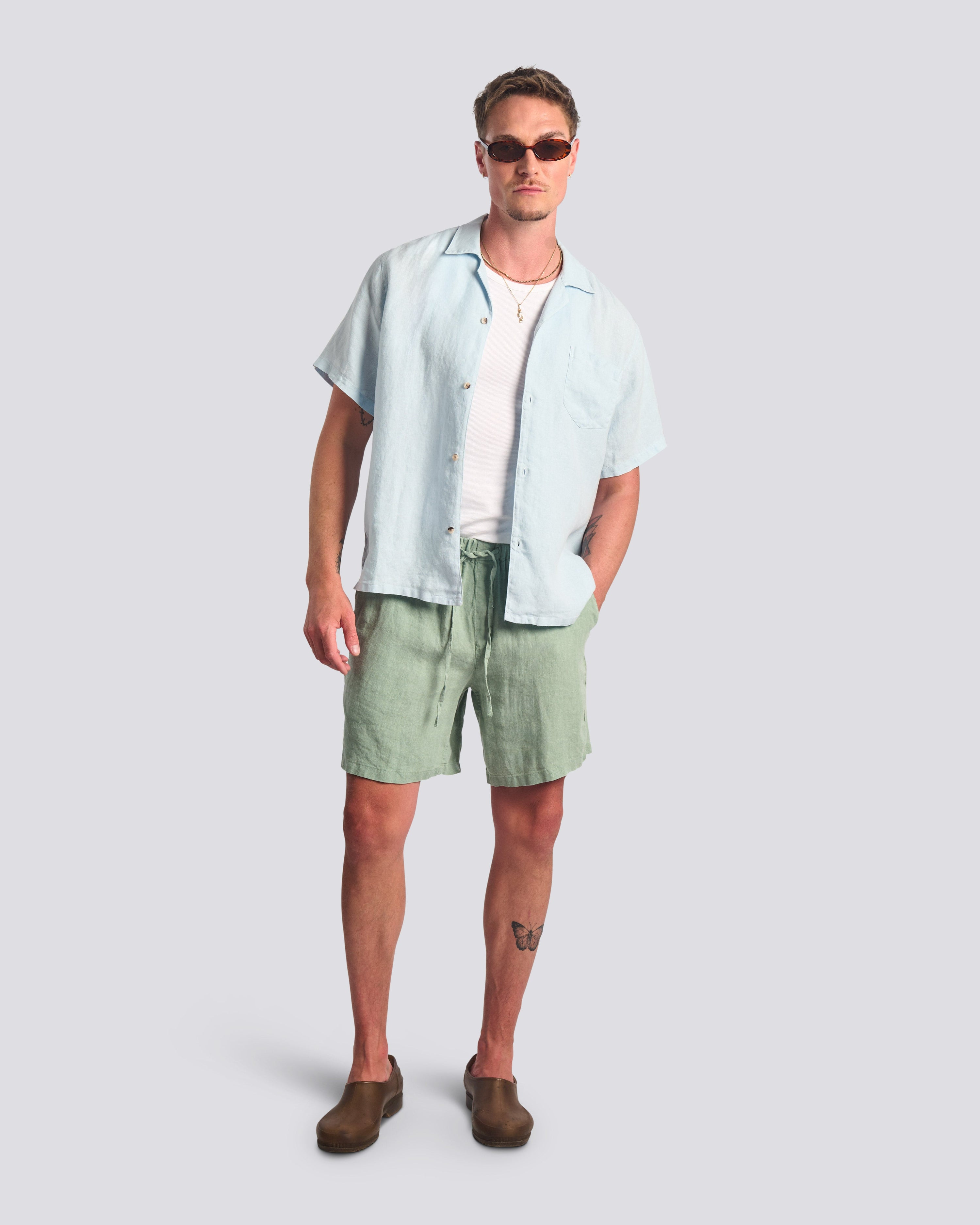 Bather Men's Sky Linen Camp Shirt in light blue, styled open over a white t-shirt with Sage linen shorts and brown loafers on a model wearing sunglasses.