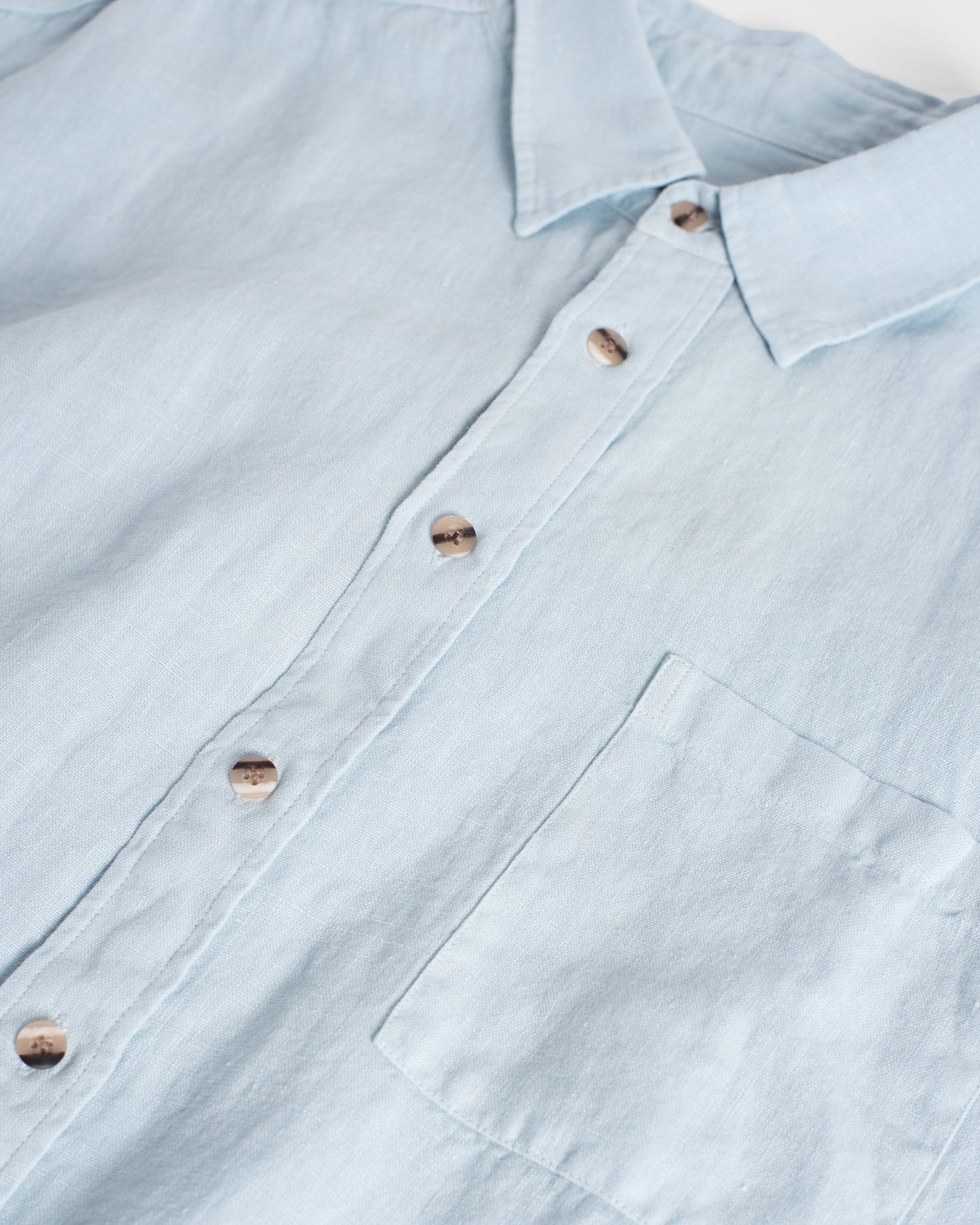Close-up of the Bather Men's Sky Linen Leisure Shirt in light blue, highlighting the textured 100% linen weave, brown mottled buttons, and the front chest pocket.