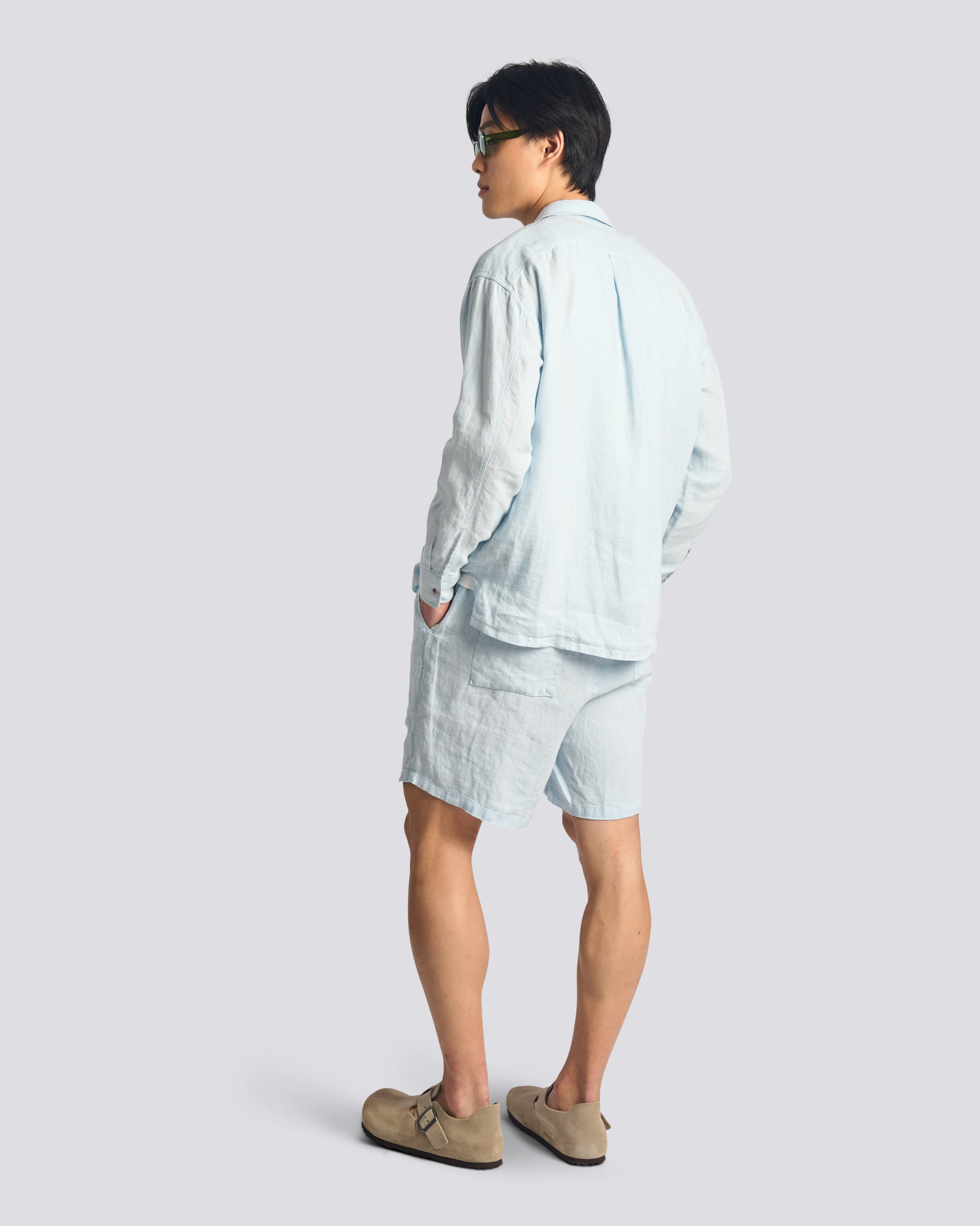 Side-rear view of the Bather Men's Sky Linen Leisure Shirt in light blue, showing the regular fit and long sleeves on a model wearing matching Sky linen shorts and tan loafers.