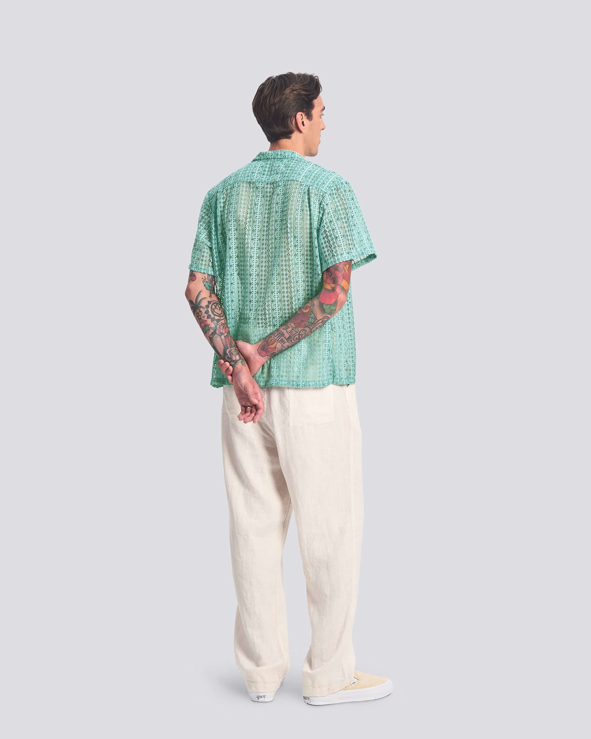 Back view of a model wearing the Bather Men's Aqua Gonzo Lace Camp Shirt, showcasing the intricate teal floral lace pattern and relaxed short-sleeve fit paired with cream trousers.