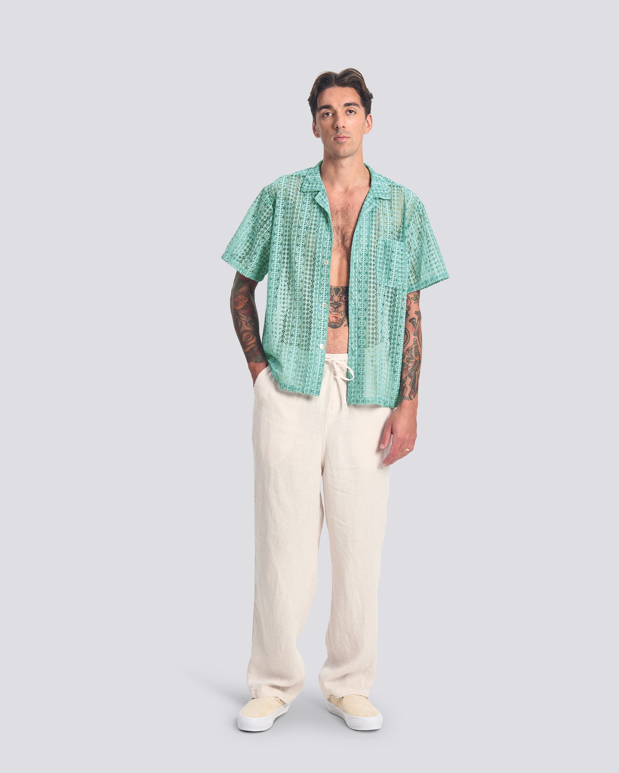 A full-length studio shot of a model wearing the Aqua Gonzo Lace Camp Shirt. The shirt features an intricate teal-colored floral lace pattern, short sleeves, and a classic camp collar. It is styled open over cream-colored linen trousers.