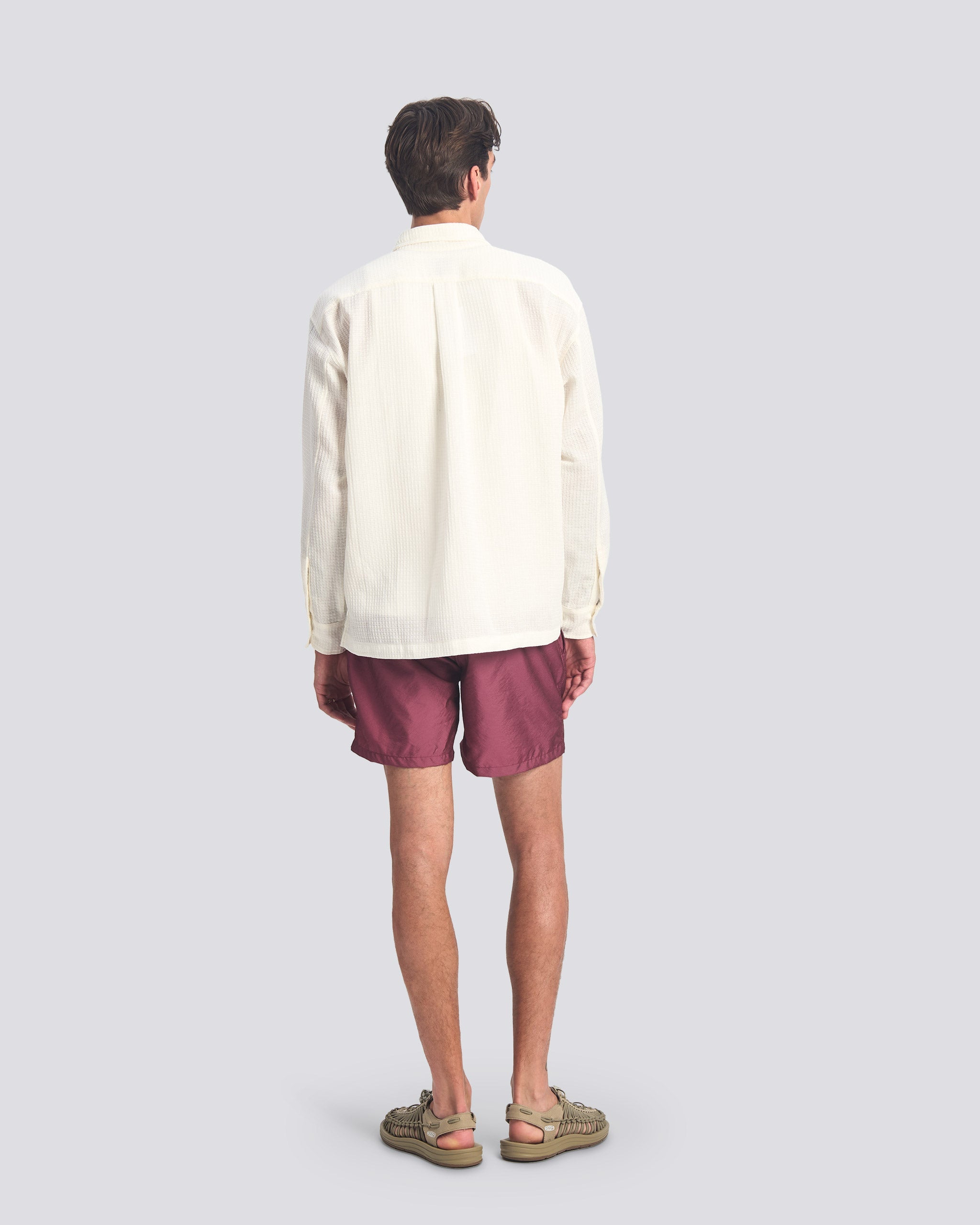 Rear view of the Bather Men's White Waffle Leisure Shirt in white, showcasing the oversized long-sleeve fit and textured cotton-linen waffle weave on a model wearing plum-colored swim trunks.
