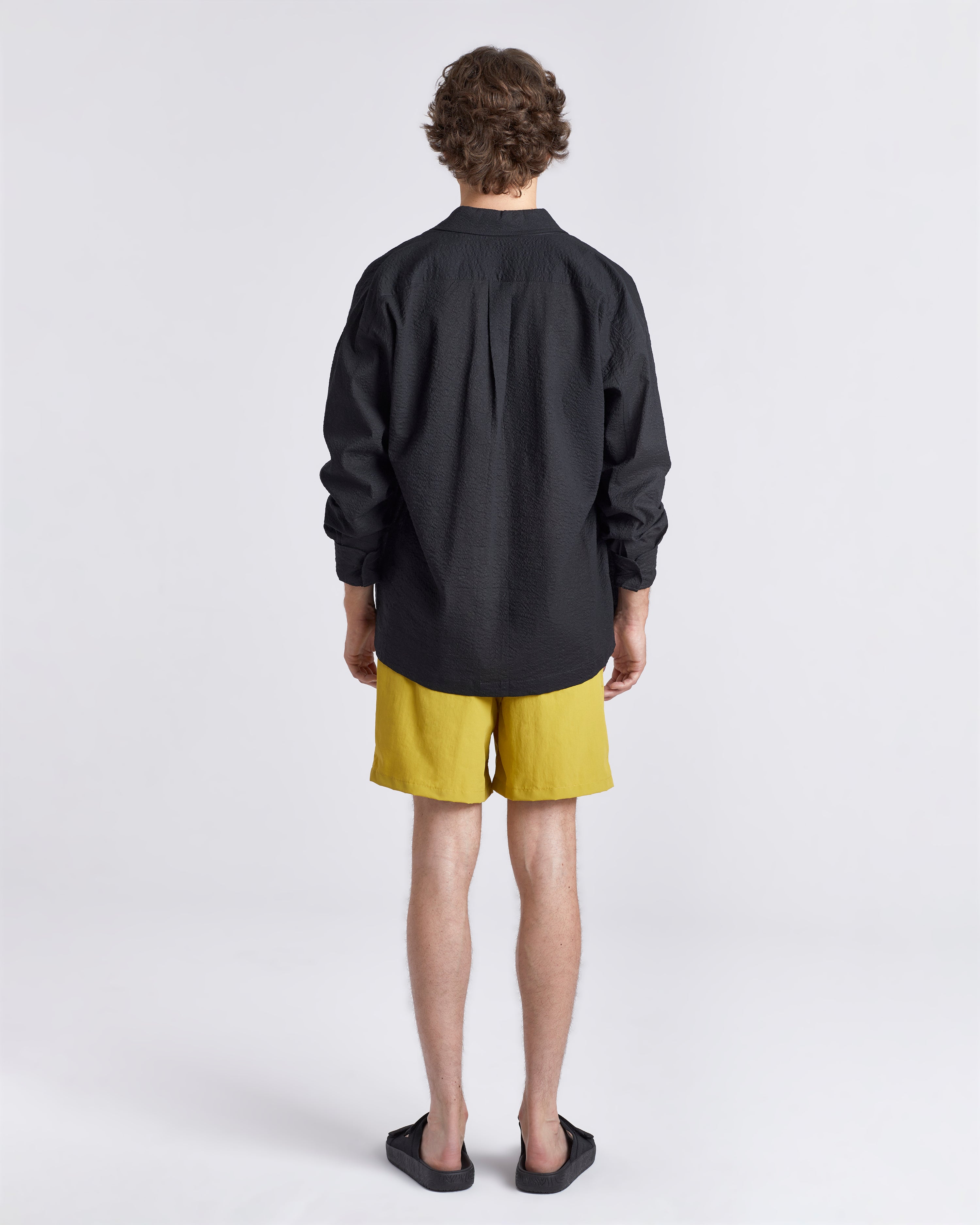 Back of man in black linen Bather leisure shirt wearing solid yellow swim trunk featuring an elastic drawstring waistband on white background