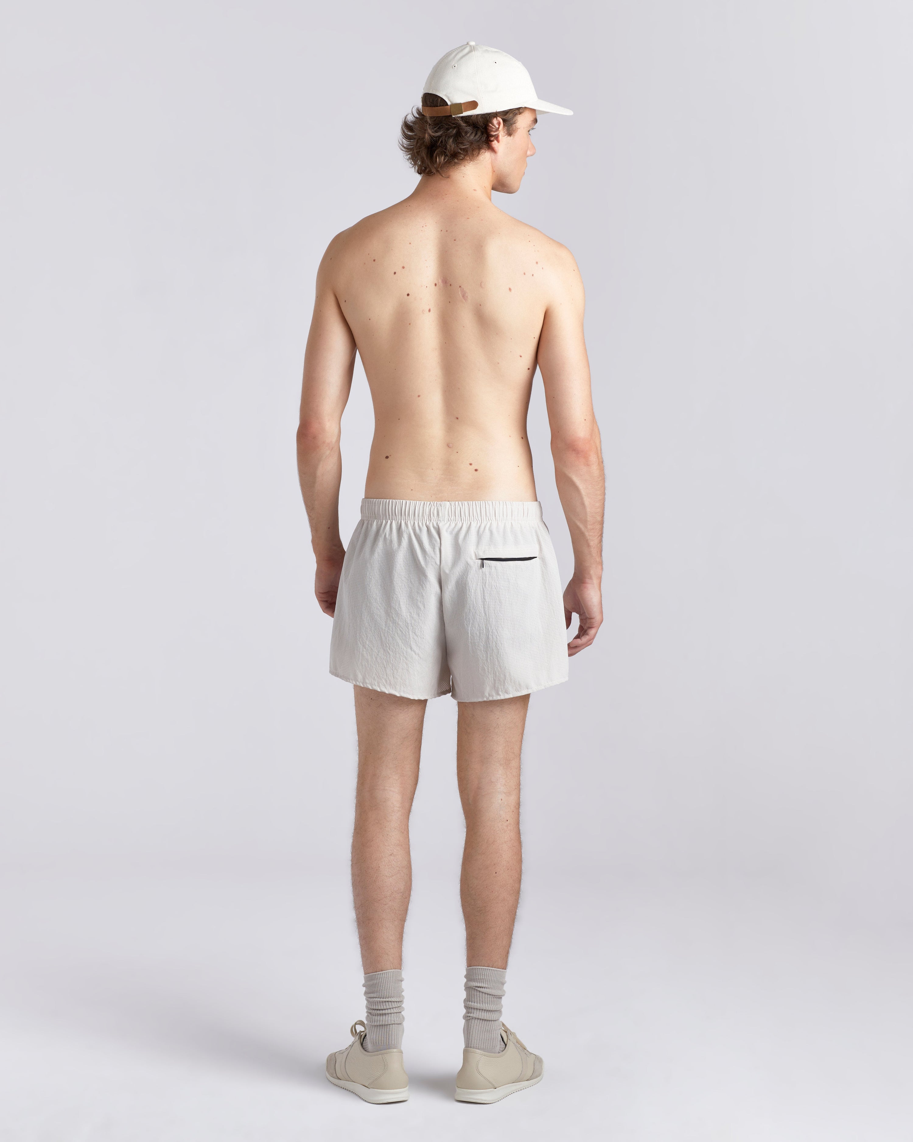 Back view of shirtless model wearing a baseball cap and 3.5 inch inseam Bather Active Trunk in white featuring lightweight recycled Japanese DotAir nylon with stretch fabric and compression liner