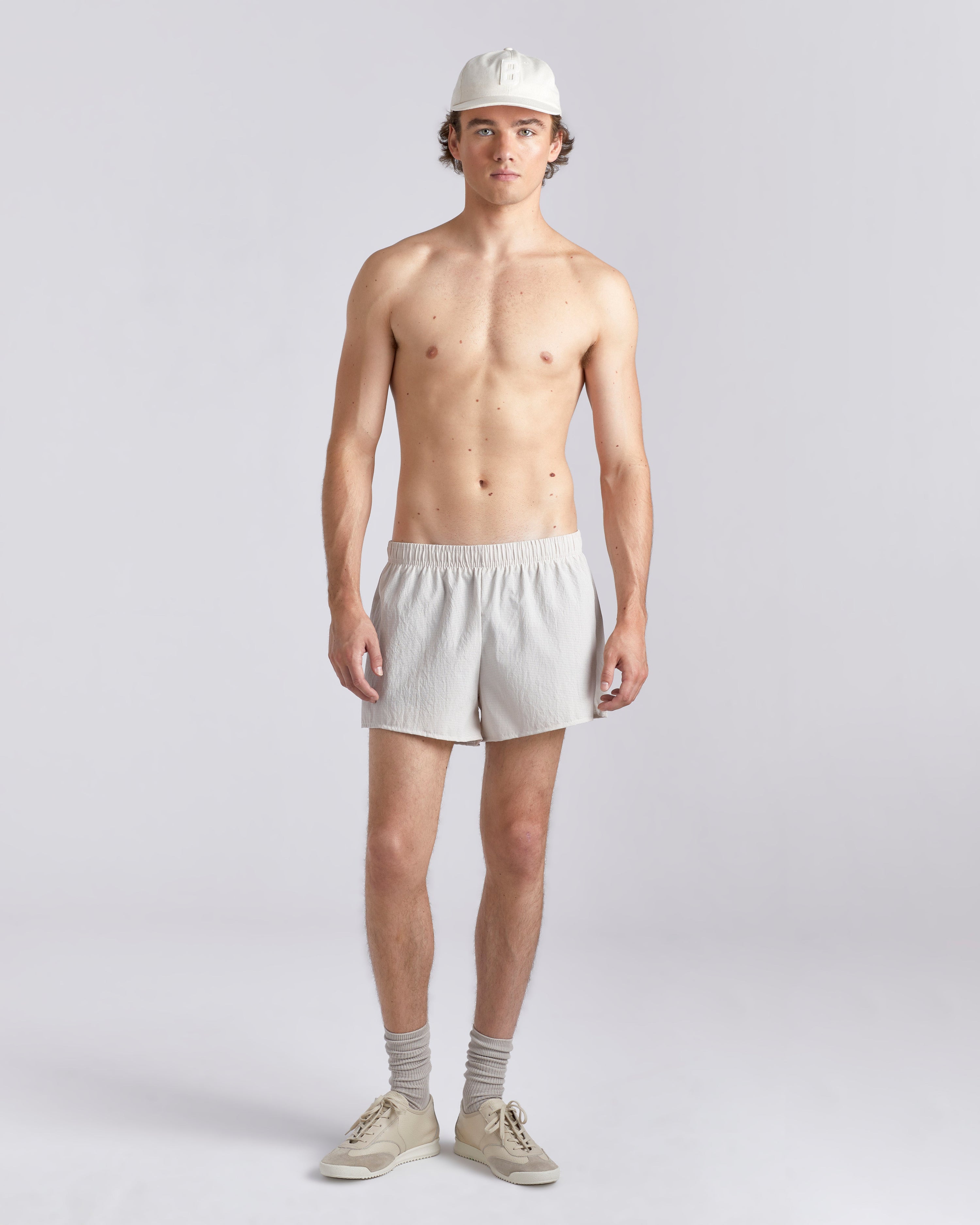 Front view of shirtless model wearing a baseball cap and 3.5 inch inseam Bather Active Trunk in white featuring lightweight recycled Japanese DotAir nylon with stretch fabric and compression liner