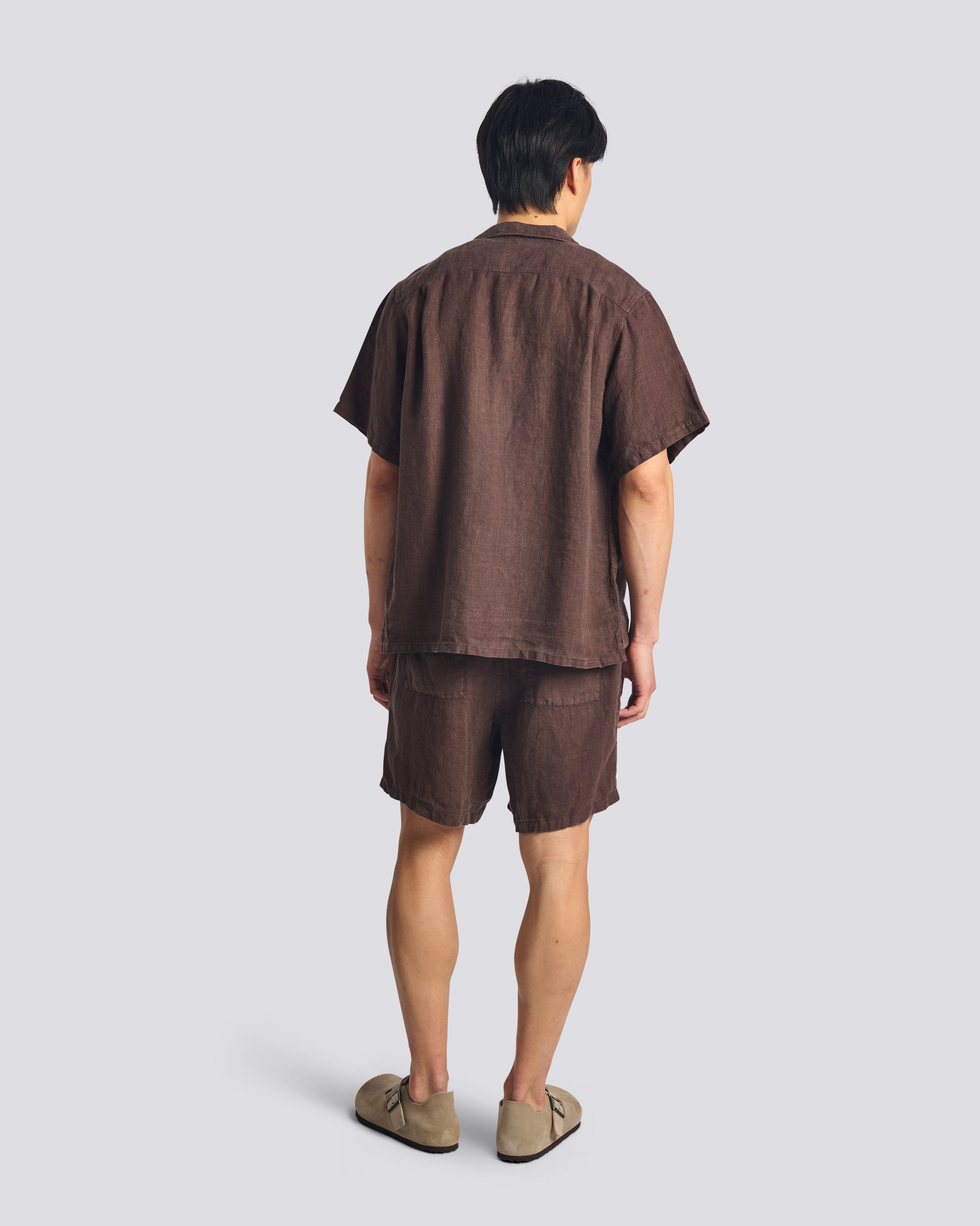 Back of male model wearing the Bather Men's Espresso Linen Camp Shirt and matching brown shorts in a relaxed fit, showcasing the lightweight drape for a classic summer vacation aesthetic.