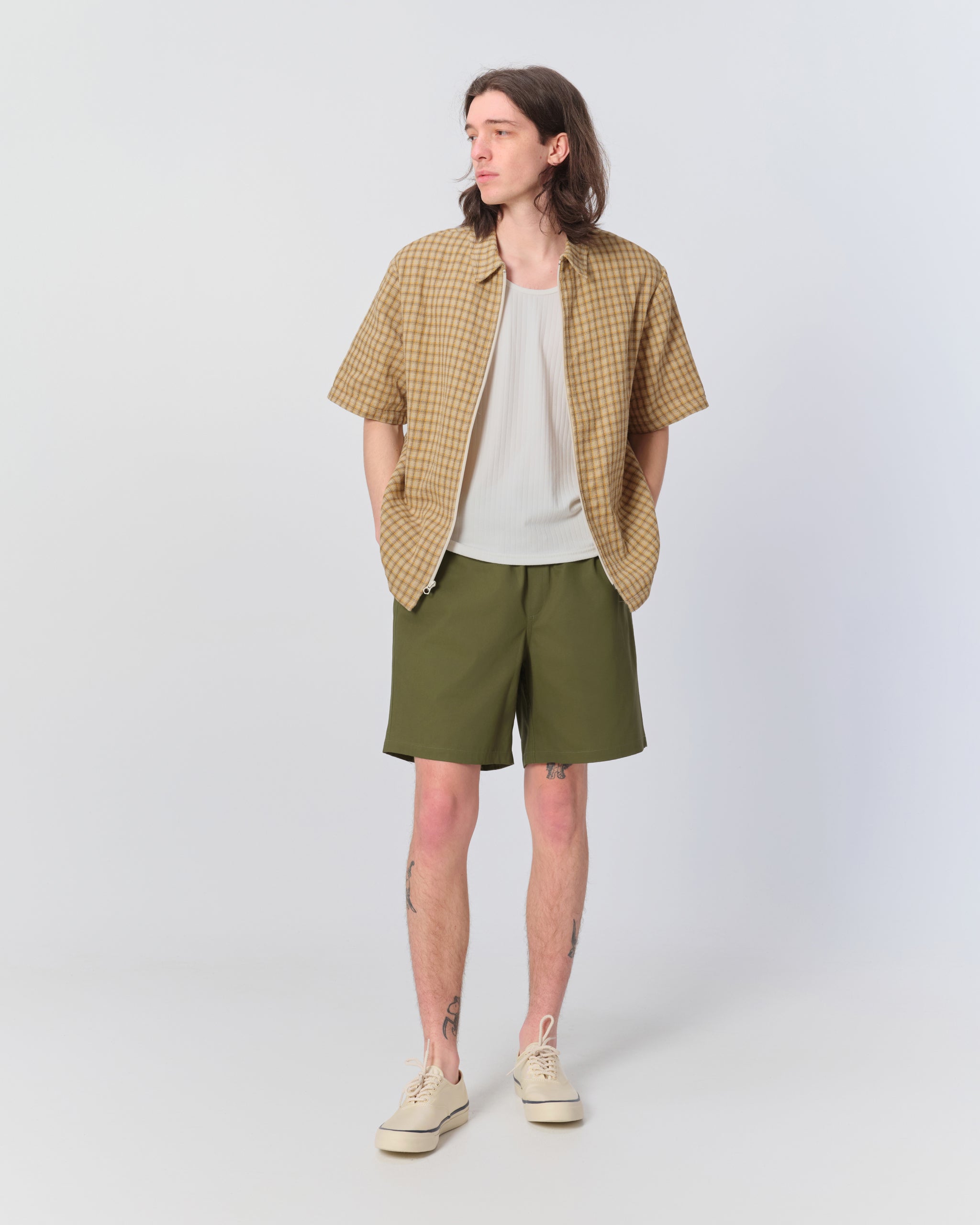 Male model wearing olive green herringbone drawstring shorts with beige checkered zip up collared shirt