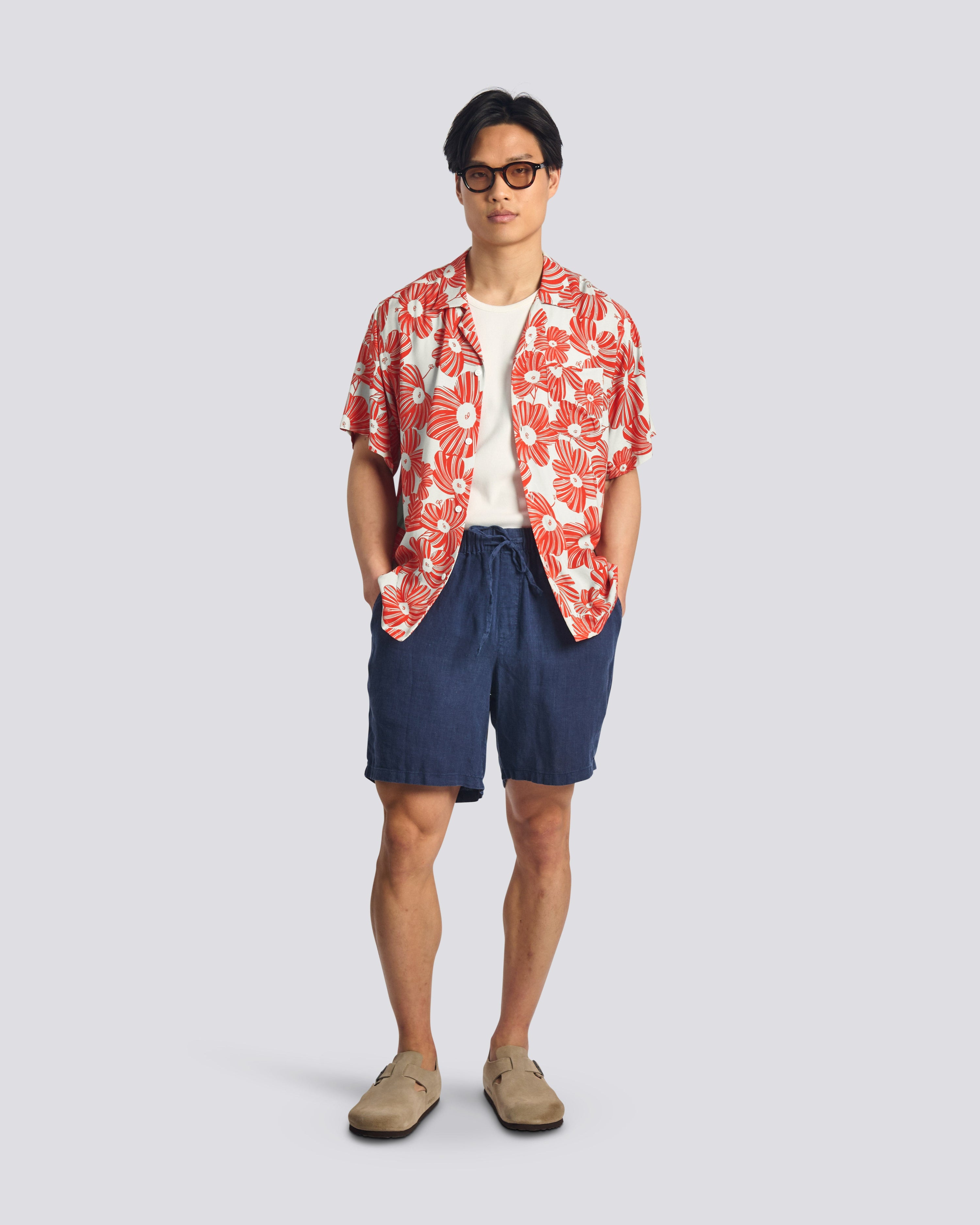 Man wearing a red floral shirt over a white t-shirt, navy shorts, and beige shoes on a light gray background
