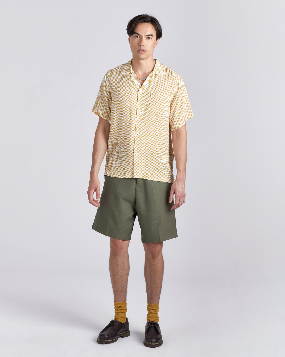A man modeling a short-sleeve button-up shirt in pale yellow with a subtle textured pattern, paired with olive green shorts, mustard yellow socks, and dark brown leather shoes, against a light gray studio background.