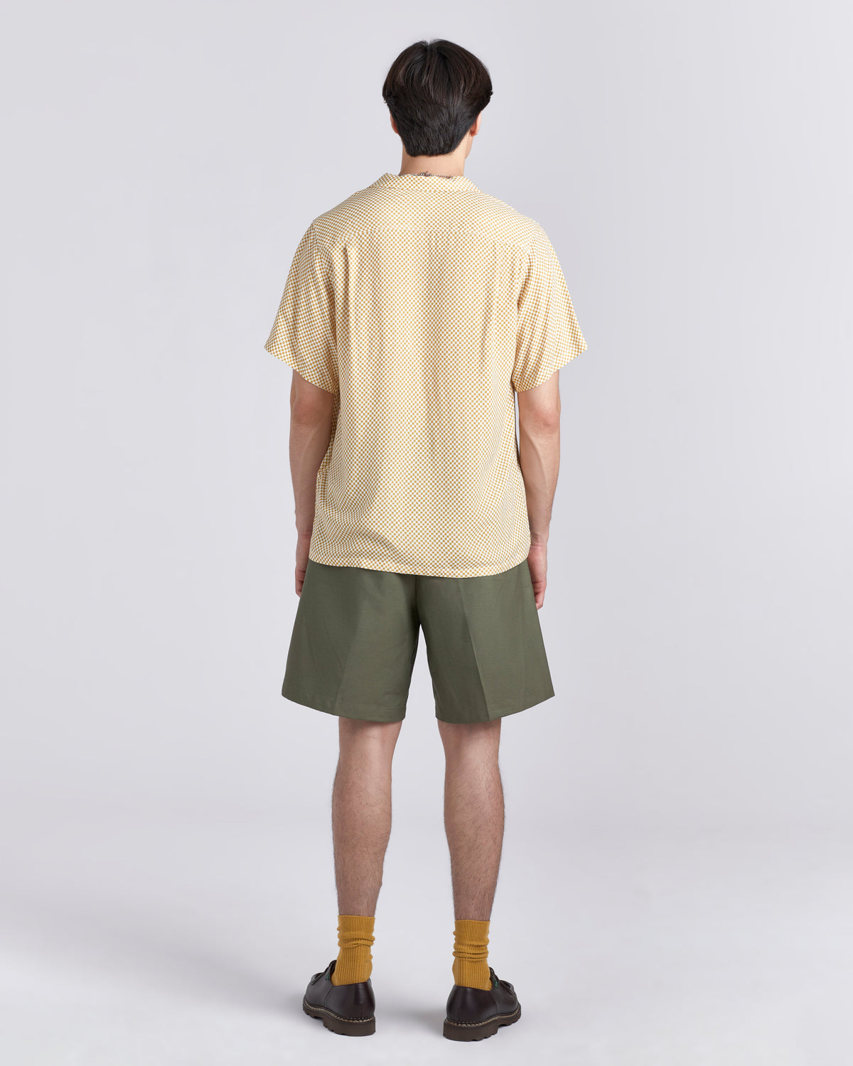 A man viewed from the back wearing a pale yellow short-sleeve textured shirt, olive green shorts, mustard yellow socks, and dark brown loafers, standing against a light gray studio background.