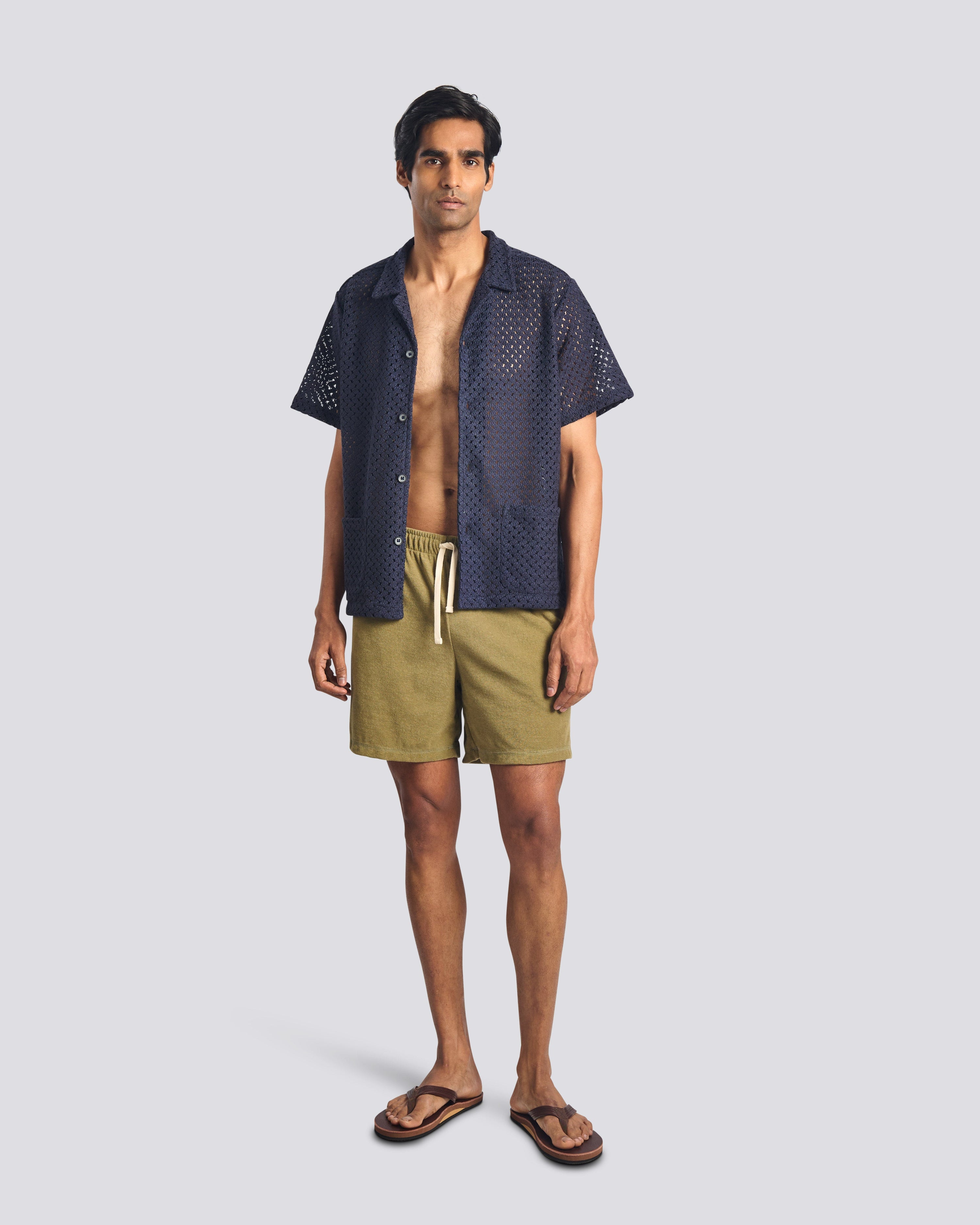 Man wearing a dark blue patterned shirt over a beige shirt and green shorts on a light gray background