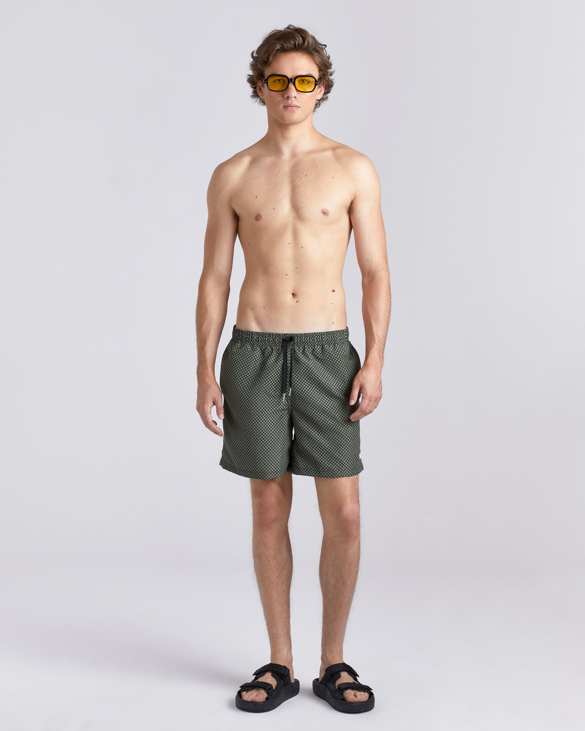 A male model stands against a light gray studio background wearing dark olive green patterned swim trunks, black sandals, and yellow-tinted sunglasses.
