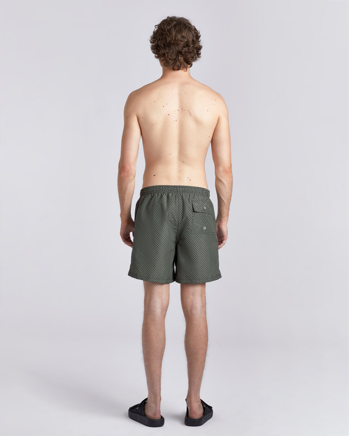 A man viewed from behind wearing dark olive green patterned swim trunks and black sandals against a light gray studio background.