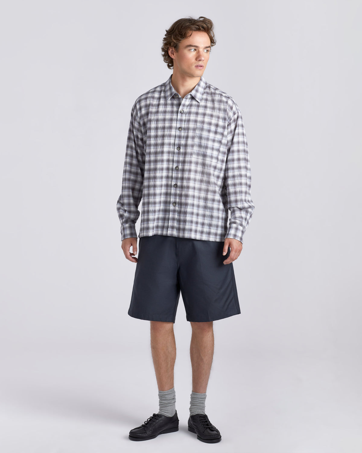 A man modeling a long-sleeve button-down plaid shirt in gray and white, paired with dark navy shorts, gray socks, and black shoes, against a light gray studio background.